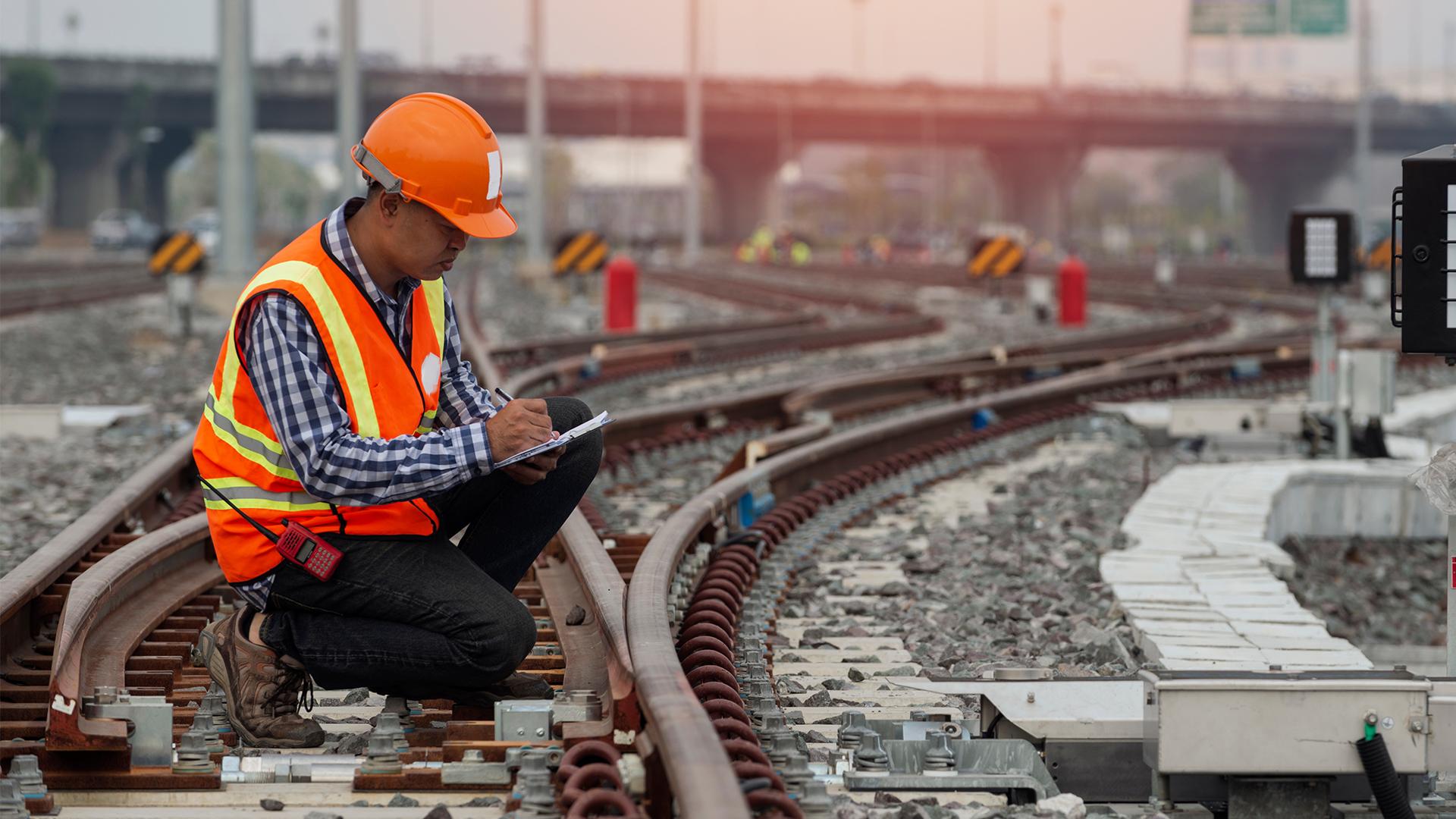 Construction worker in an orange vest and helmet kneeling on railway tracks, taking notes on a clipboard, with a bridge in the background.