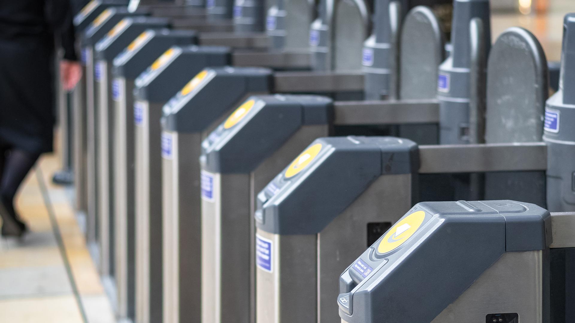 A row of electronic ticket barriers at a train station, with a person walking through the open gate on the left.