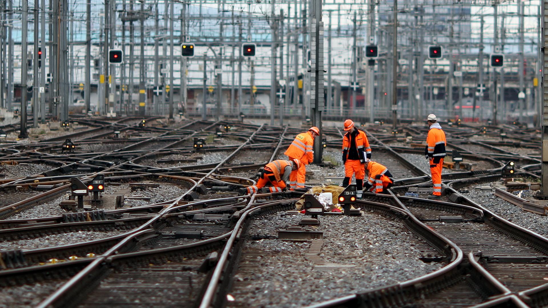 Railway workers in orange uniforms perform maintenance on a complex network of tracks, surrounded by signal lights and overhead wires.