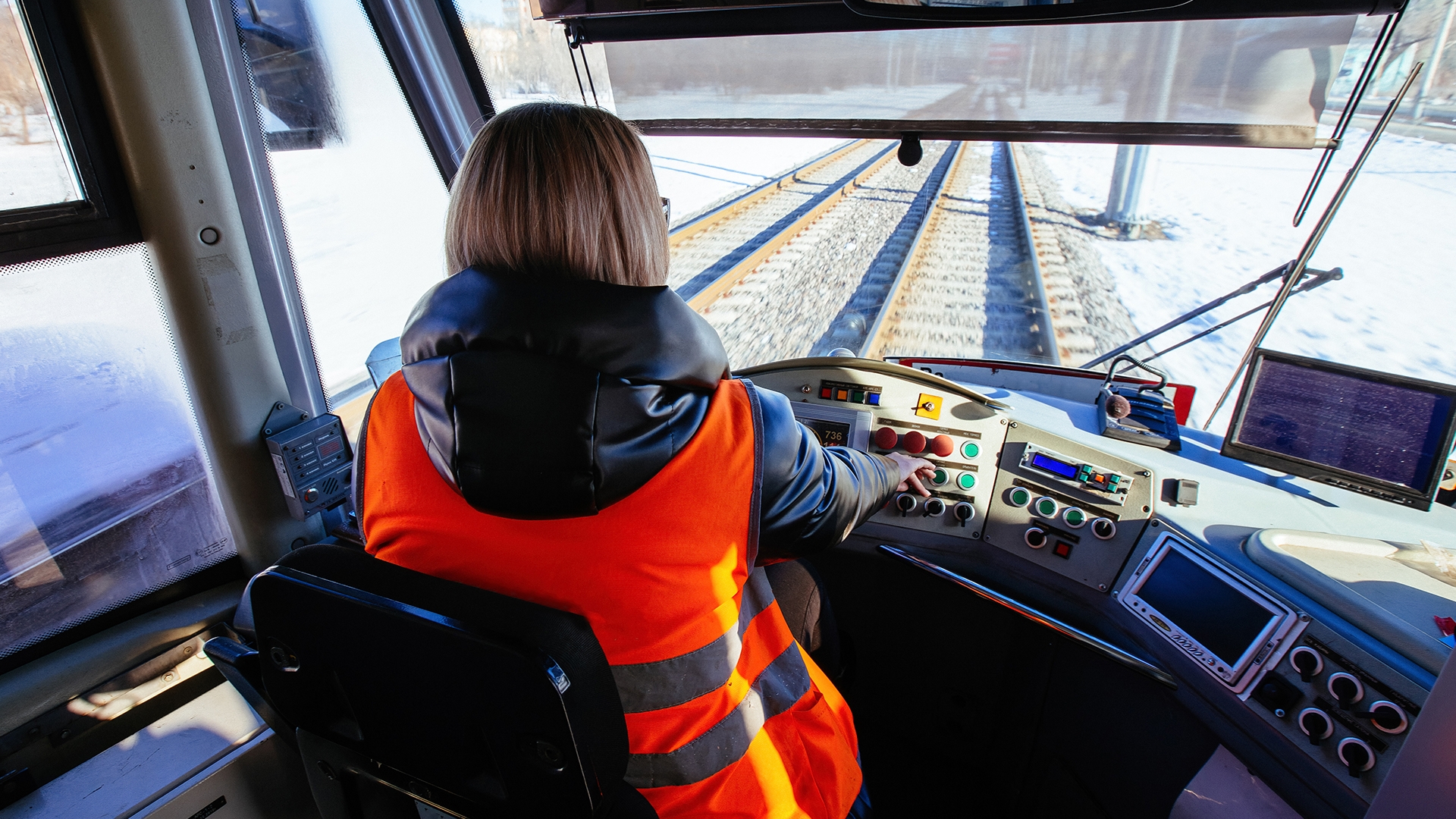 Train driver in an orange vest operates controls, navigating snowy tracks. View from behind the driver, with screens and buttons visible.