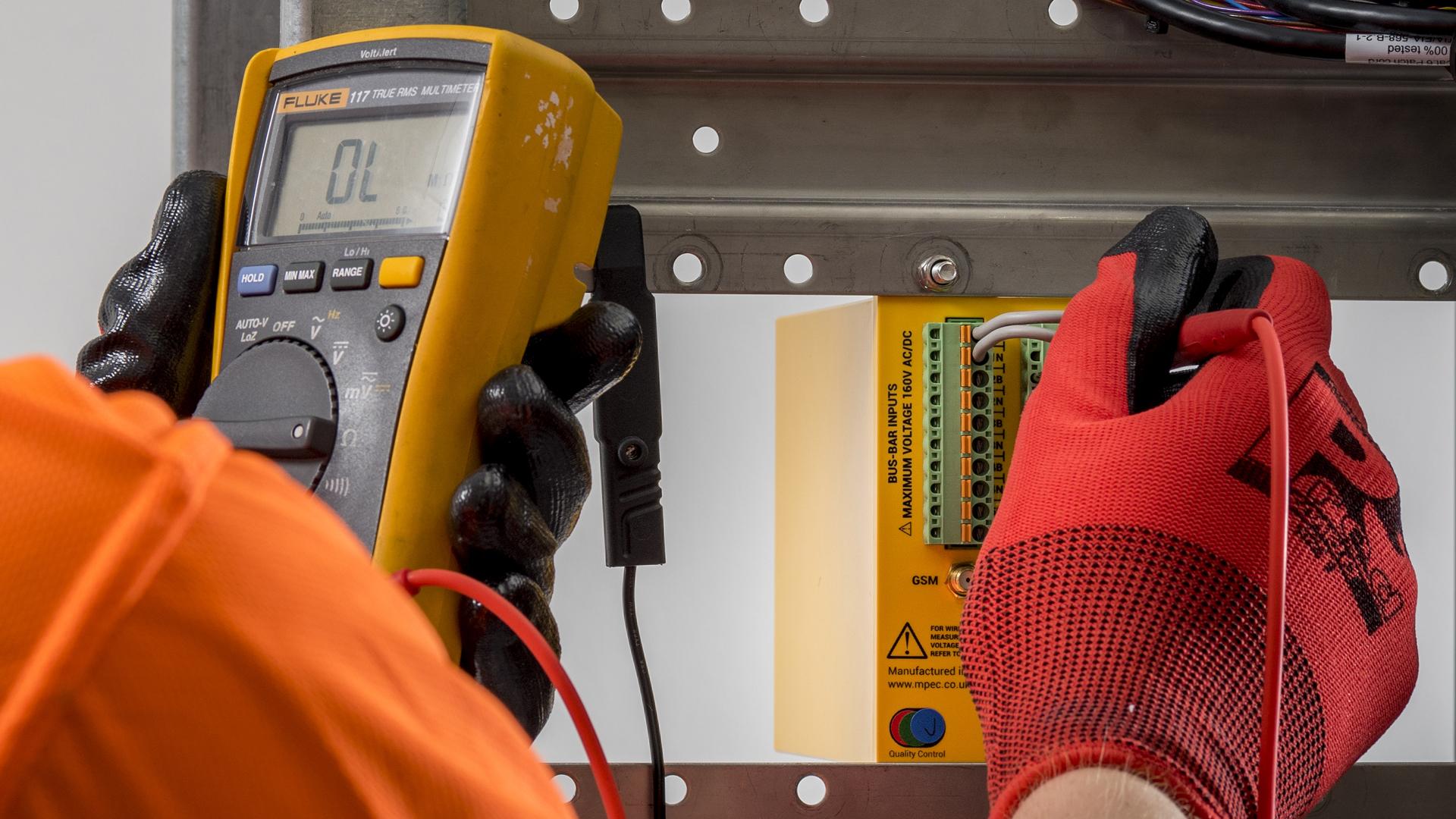 A person in orange gloves uses a digital multimeter to test an electrical device mounted on a perforated metal panel.