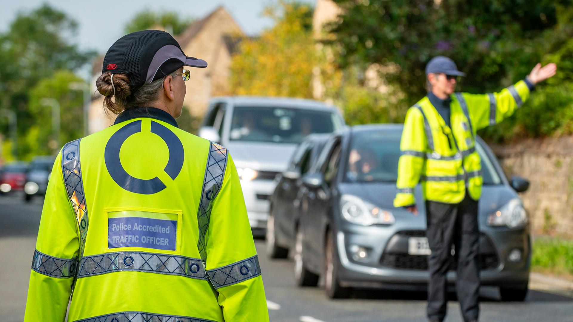Two traffic officers in high-visibility jackets manage traffic on a road. One directs vehicles while the other observes.
