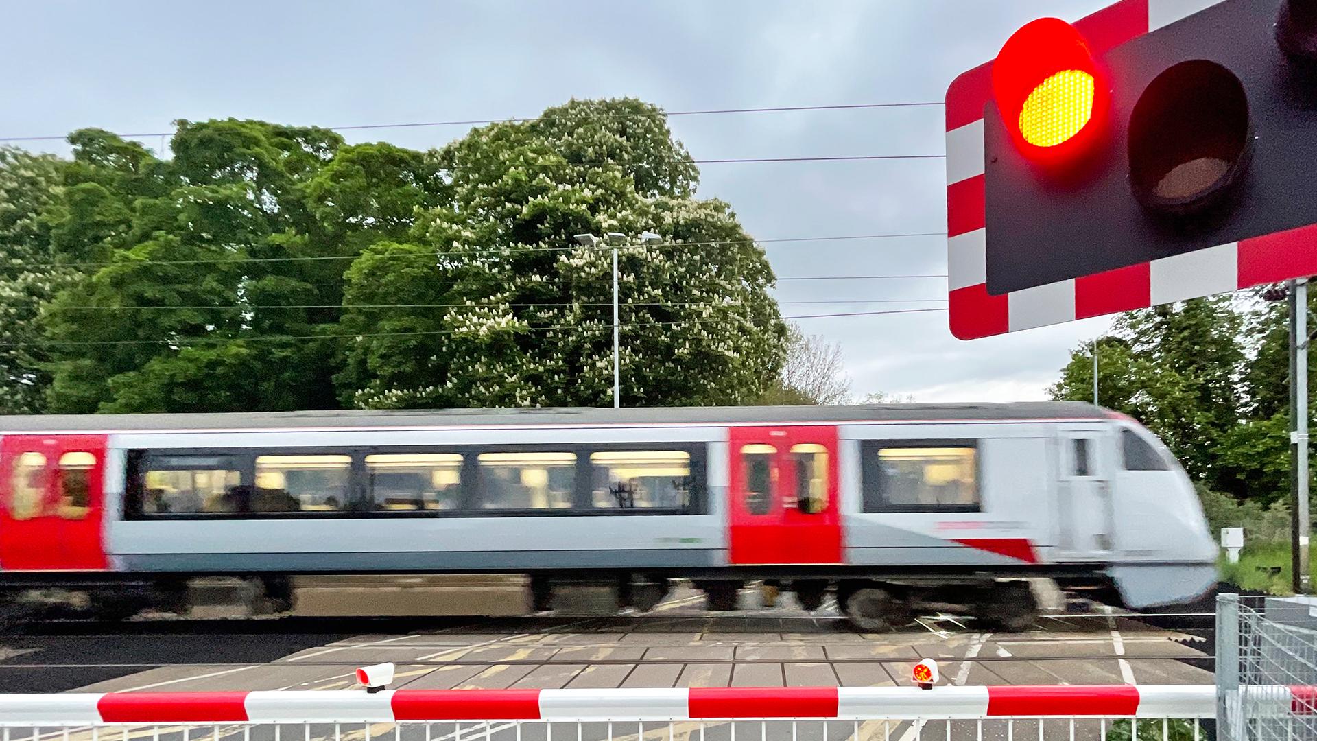 A train speeds past a railway crossing with red barriers down and a red signal light. Trees are visible in the background.