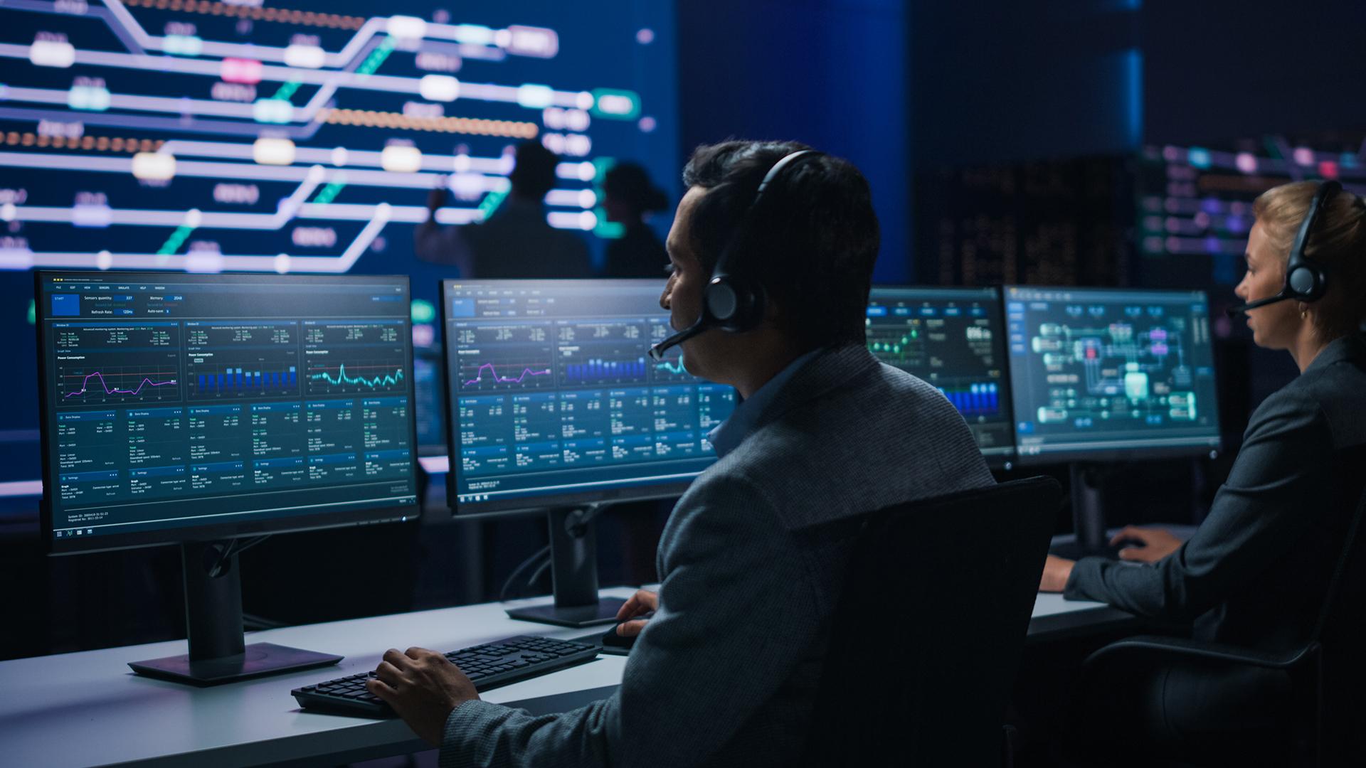 People in a control room wearing headsets, monitoring data on multiple computer screens displaying graphs and charts.