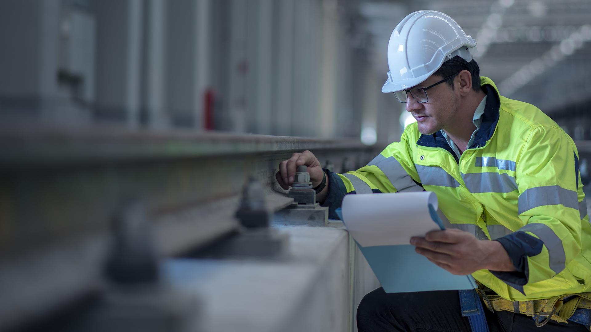 A construction worker in a high-visibility jacket and hard hat inspects railway tracks, holding a clipboard and adjusting a bolt.