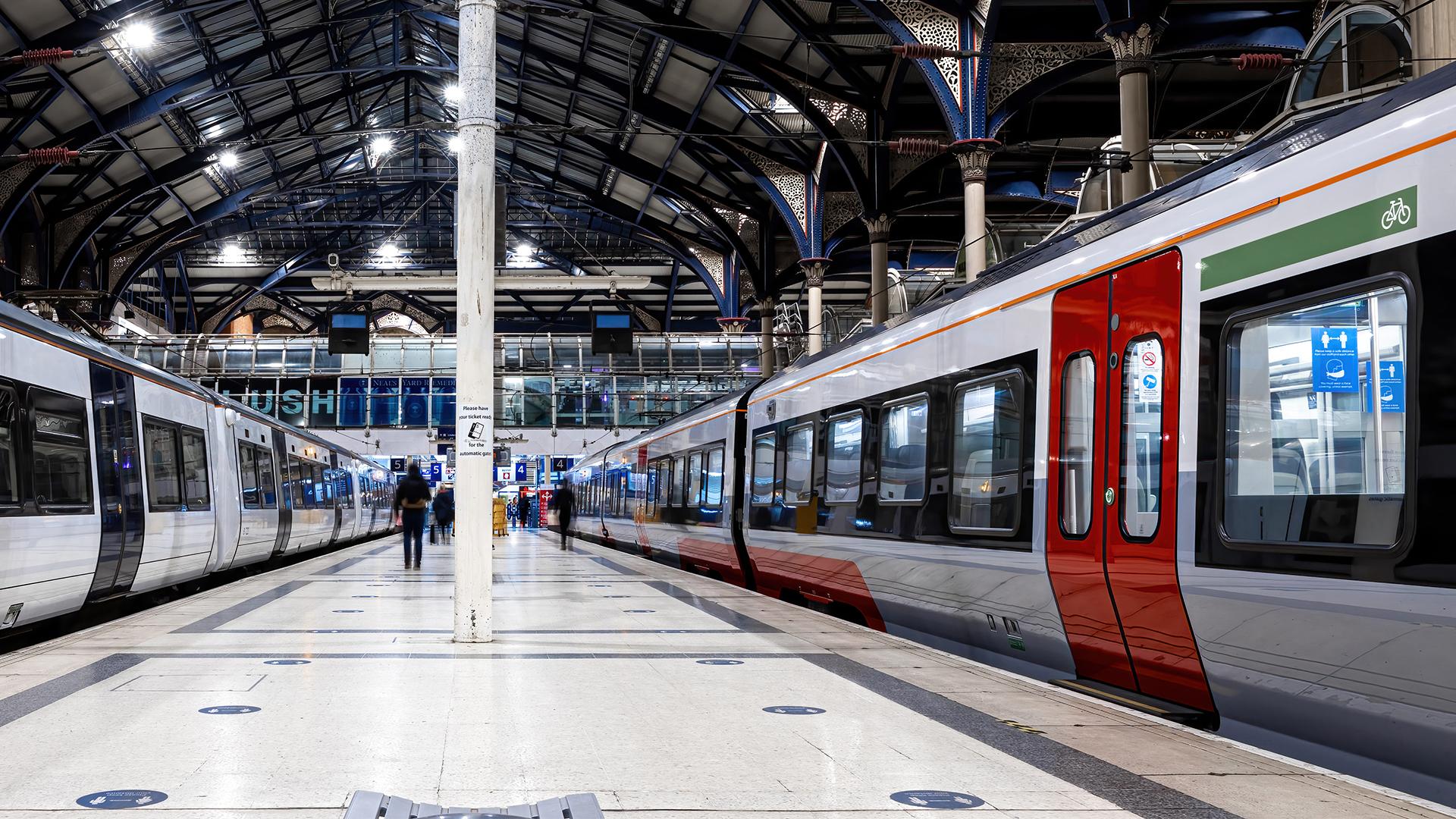 A modern train station with two trains on either side of a platform. The arched roof features metal and glass, and a few passengers are walking.
