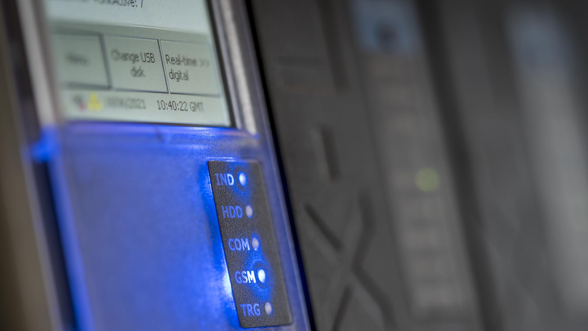 Close-up of a device with a digital display and illuminated indicator lights labeled HDD, COM, GSM, and TRG, against a blurred background.