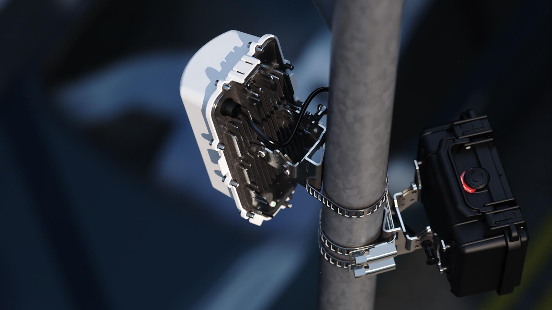 A close-up of electronic equipment mounted on a pole, featuring a white device with cables and a black box secured with metal brackets.