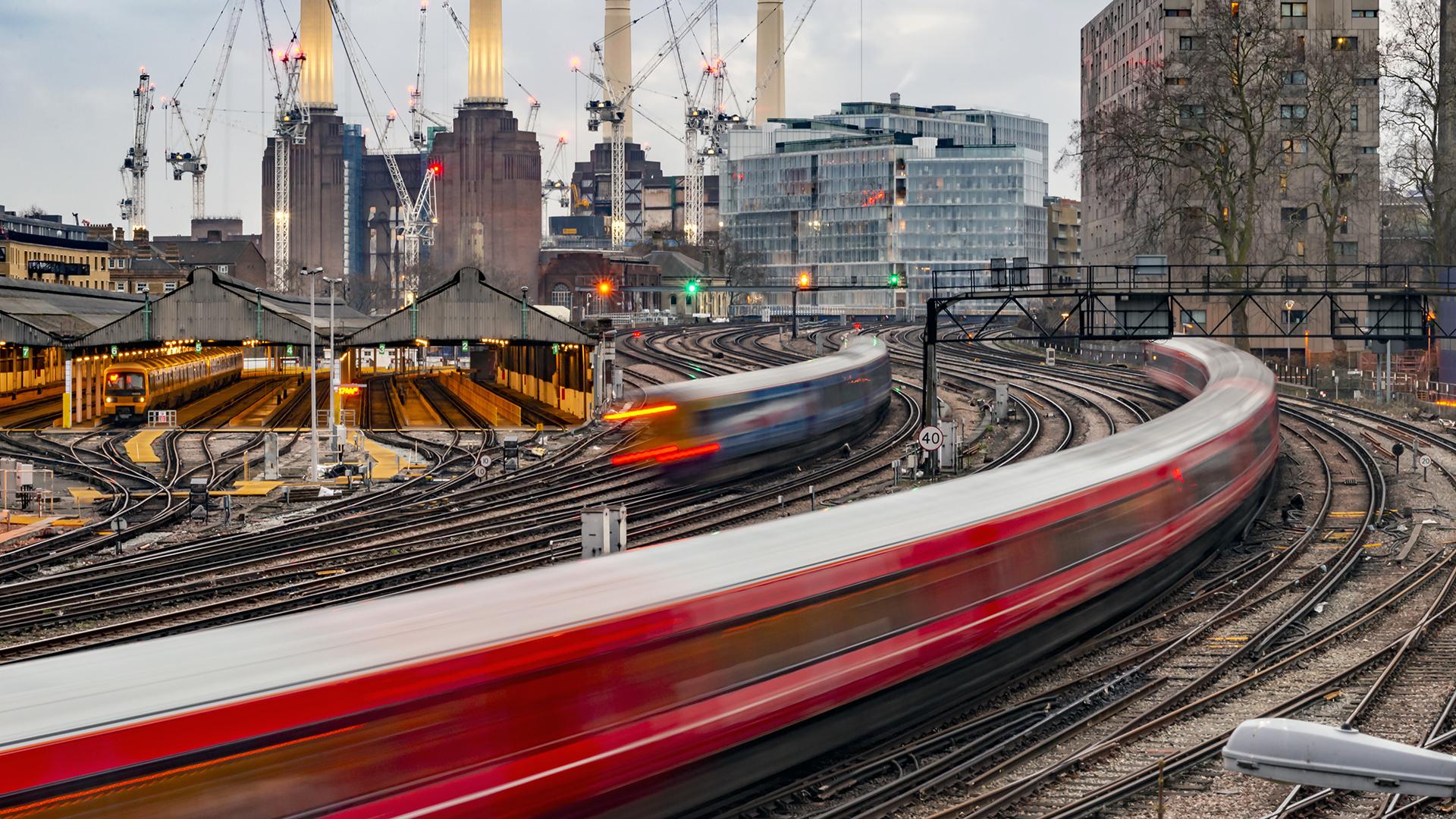 Blurred trains speed through a complex railway network near an urban station, with tall buildings and cranes in the background.