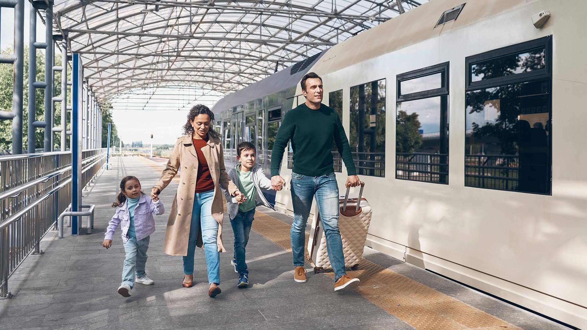 Family with two children and a toddler walking on a train platform, holding hands, under a glass canopy.