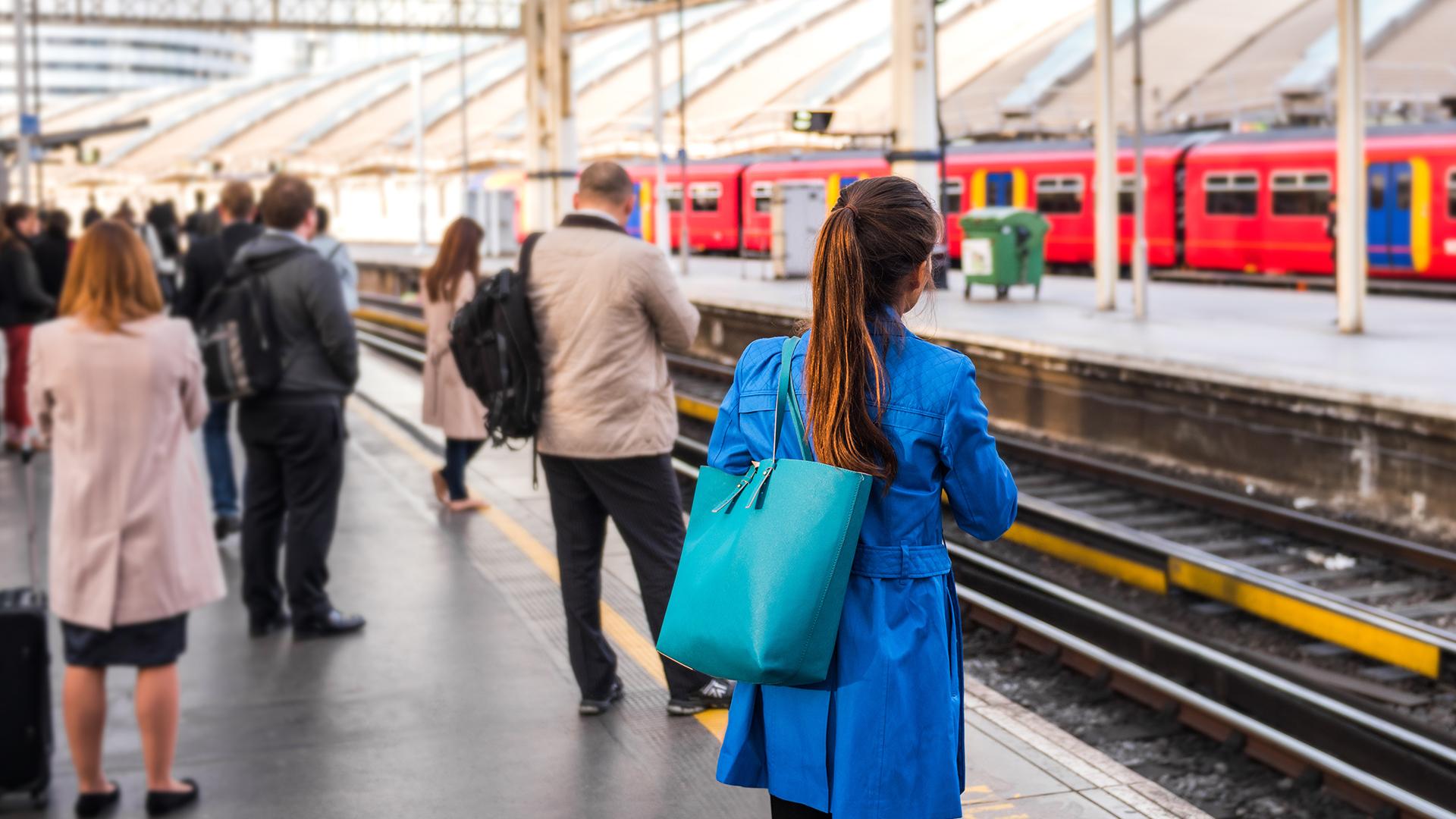 People waiting on a train platform, with a woman in a blue coat in the foreground and a red train approaching in the background.