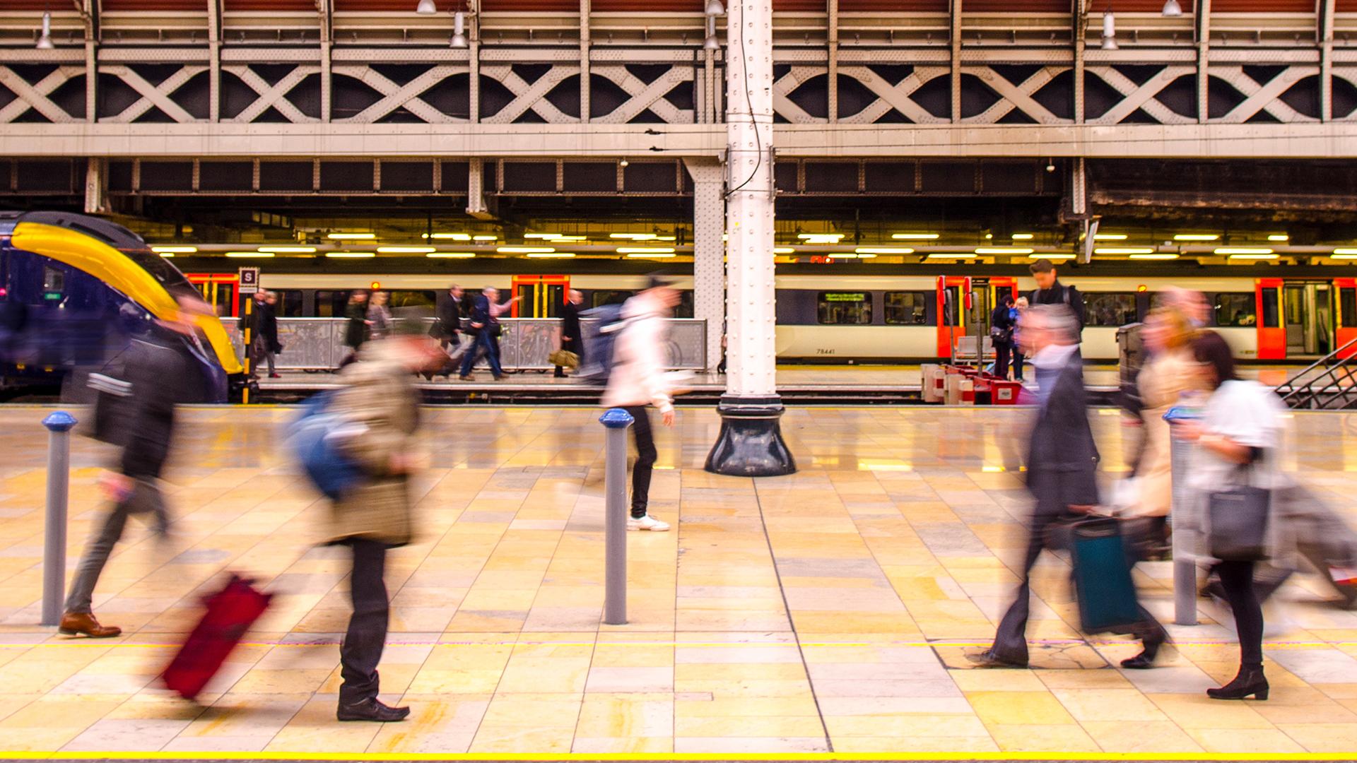 Blurred motion of commuters with luggage walking in a busy train station, with trains visible in the background.