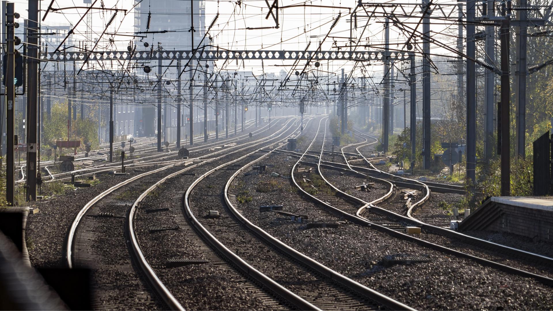 Multiple railway tracks curve into the distance under a network of overhead power lines, with a misty urban backdrop.