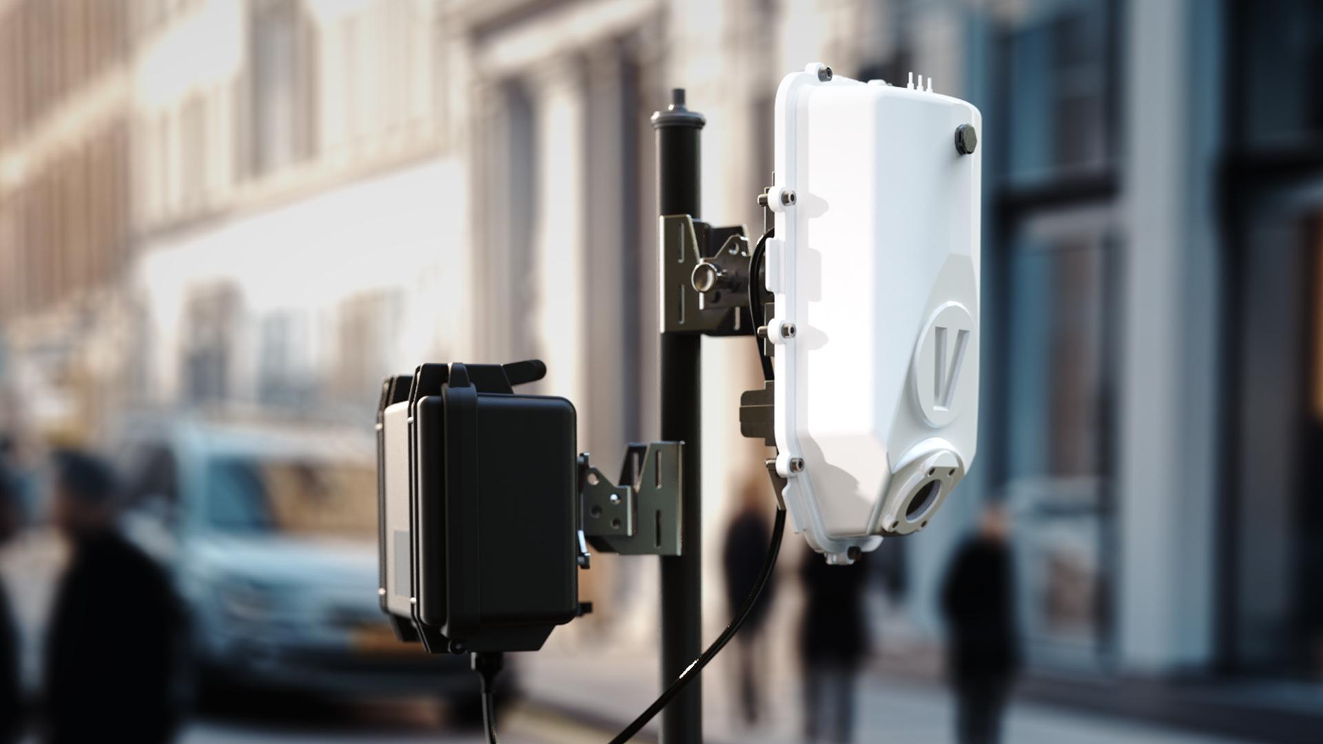 Close-up of a 5G small cell antenna mounted on a city street pole, with blurred pedestrians and buildings in the background.