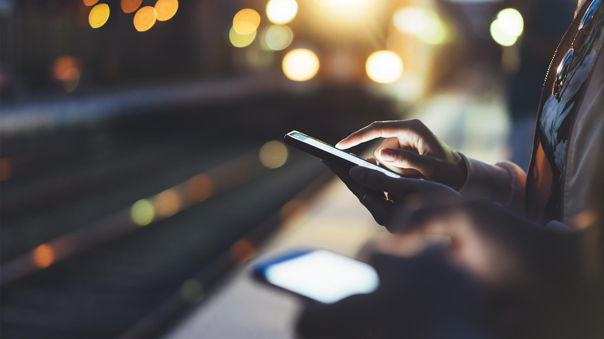 People using smartphones at a dimly lit train station, with blurred lights in the background creating a warm ambiance.
