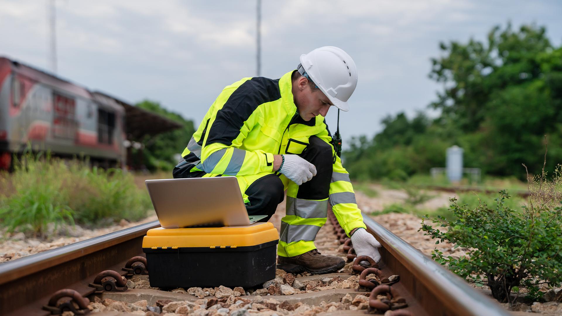 Worker in high-visibility gear and helmet inspects railway tracks using a laptop on a toolbox. Train and greenery in the background.