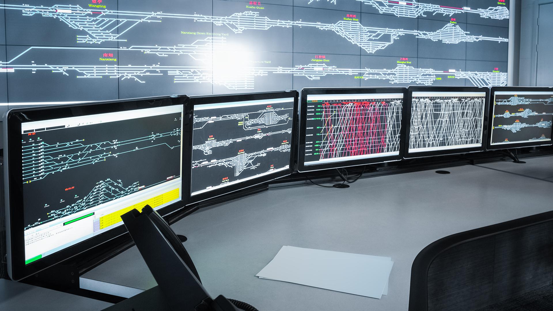 Control room with multiple monitors displaying complex railway track diagrams and data, against a large wall screen with similar schematics.
