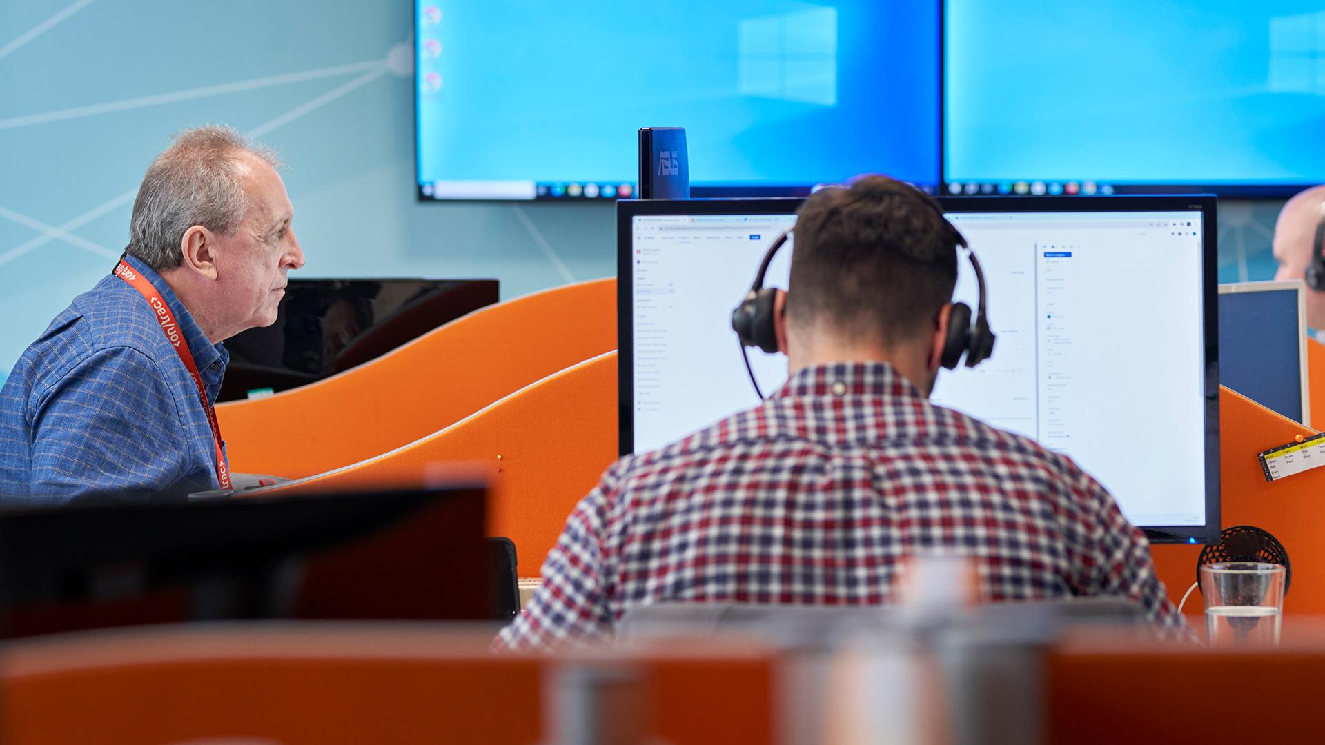 Two men working at desks with computers in an office. One wears headphones, and the other looks at a screen. Bright orange partitions separate them.