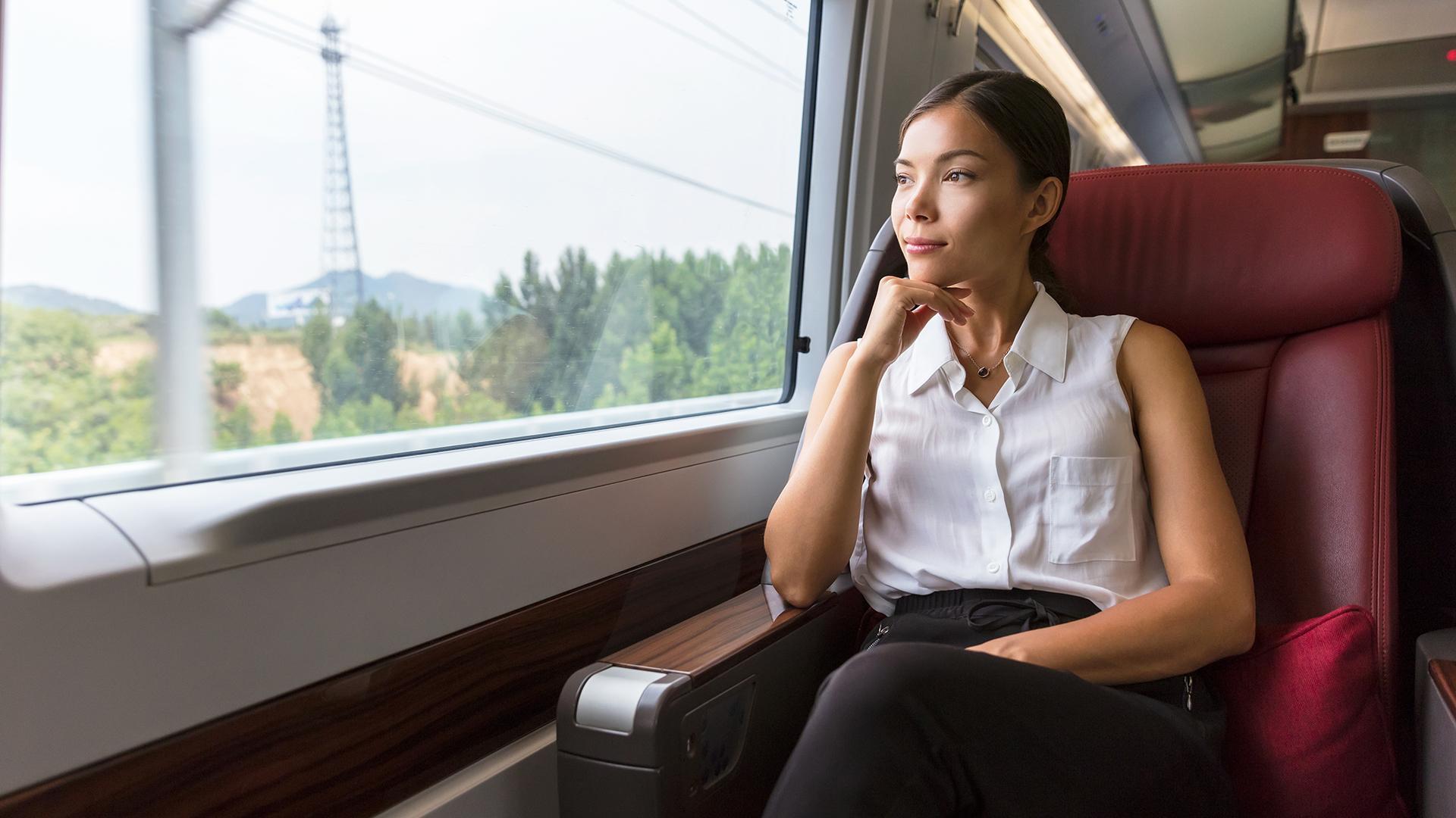 Woman in a white blouse sitting on a train, gazing out the window at passing trees and distant hills, with a thoughtful expression.