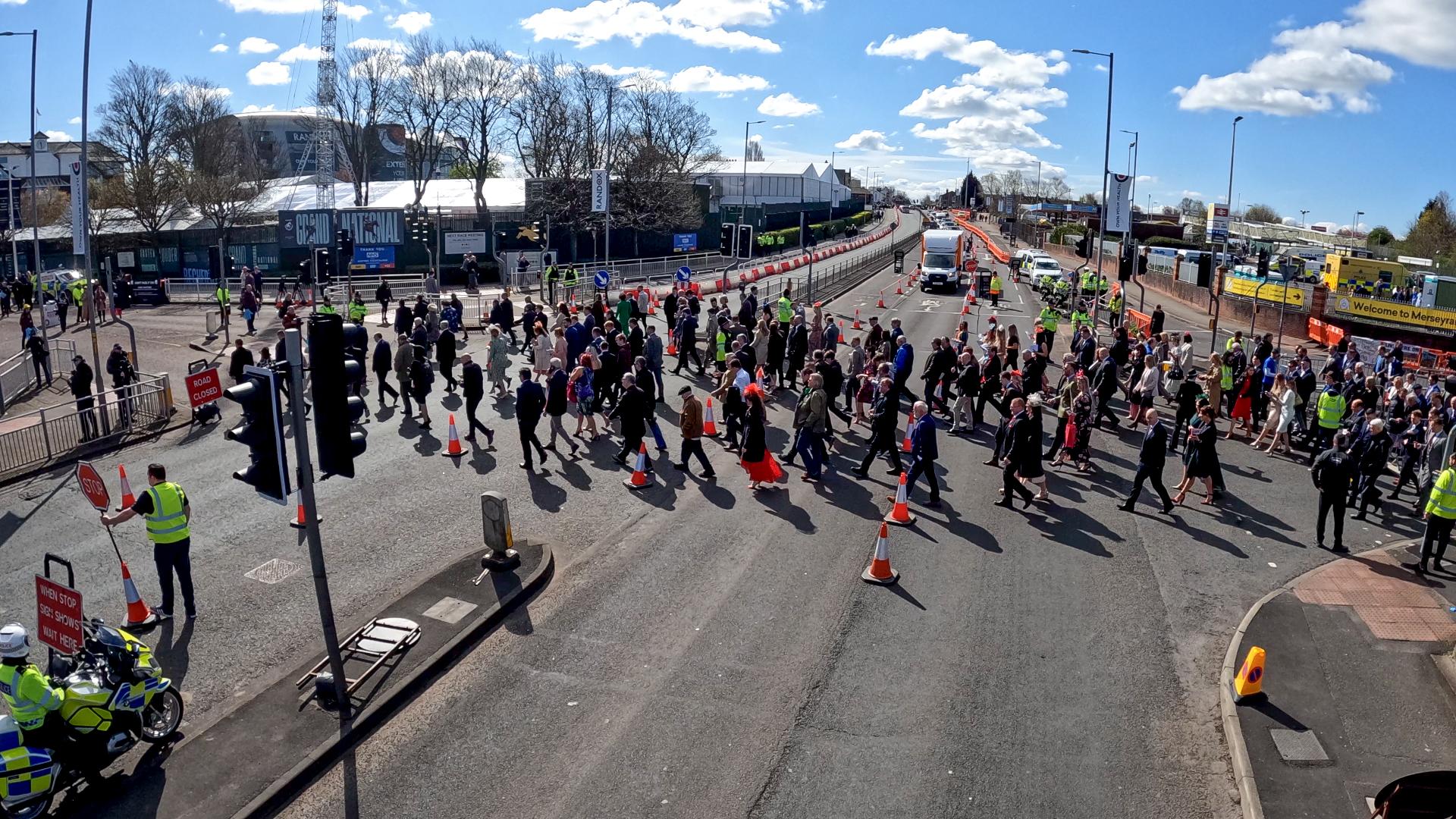 A large crowd of people crossing a street with traffic cones and police officers managing the scene under a clear blue sky.