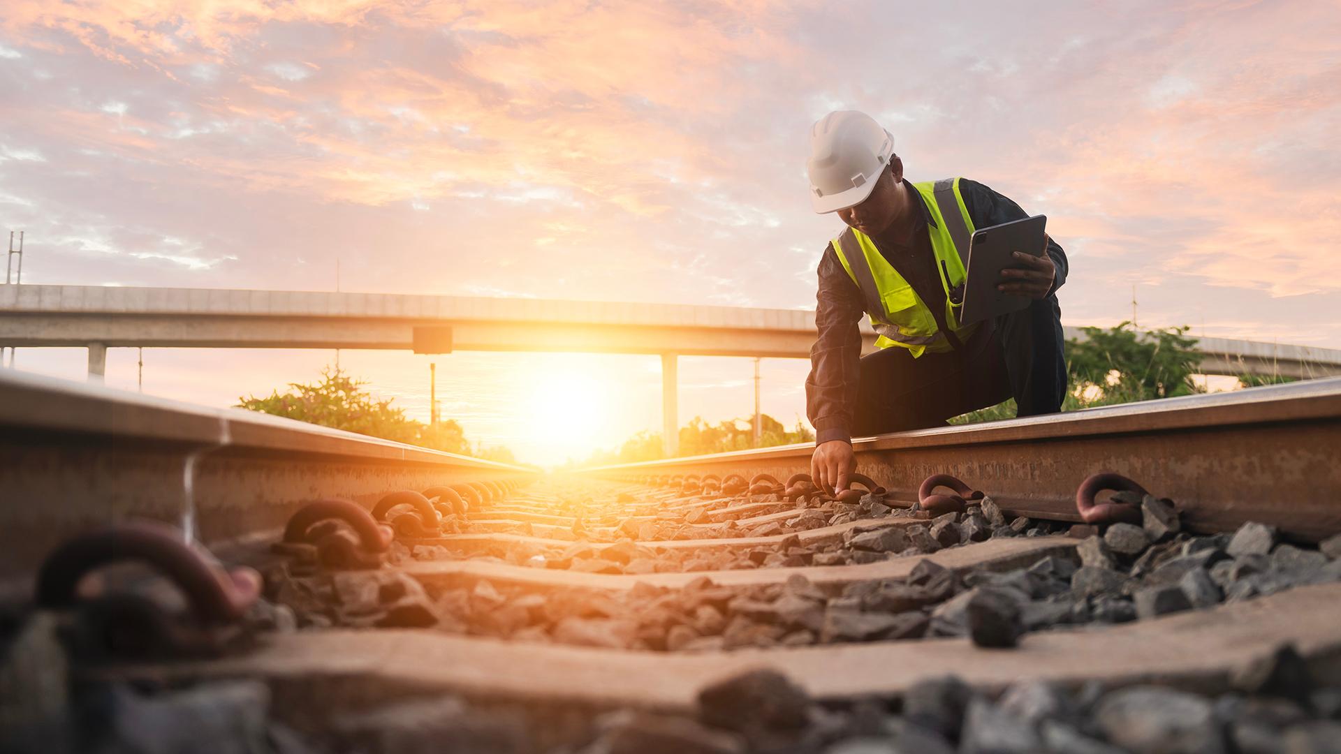 Worker in safety gear inspects railway tracks at sunset, holding a tablet. Elevated highways and a vibrant sky are in the background.