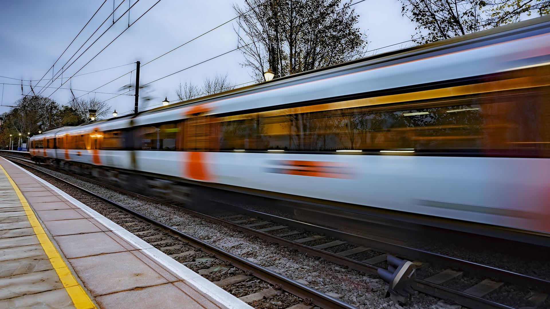 A fast-moving train blurs past a station platform lined with yellow safety markings, surrounded by trees under a cloudy sky.