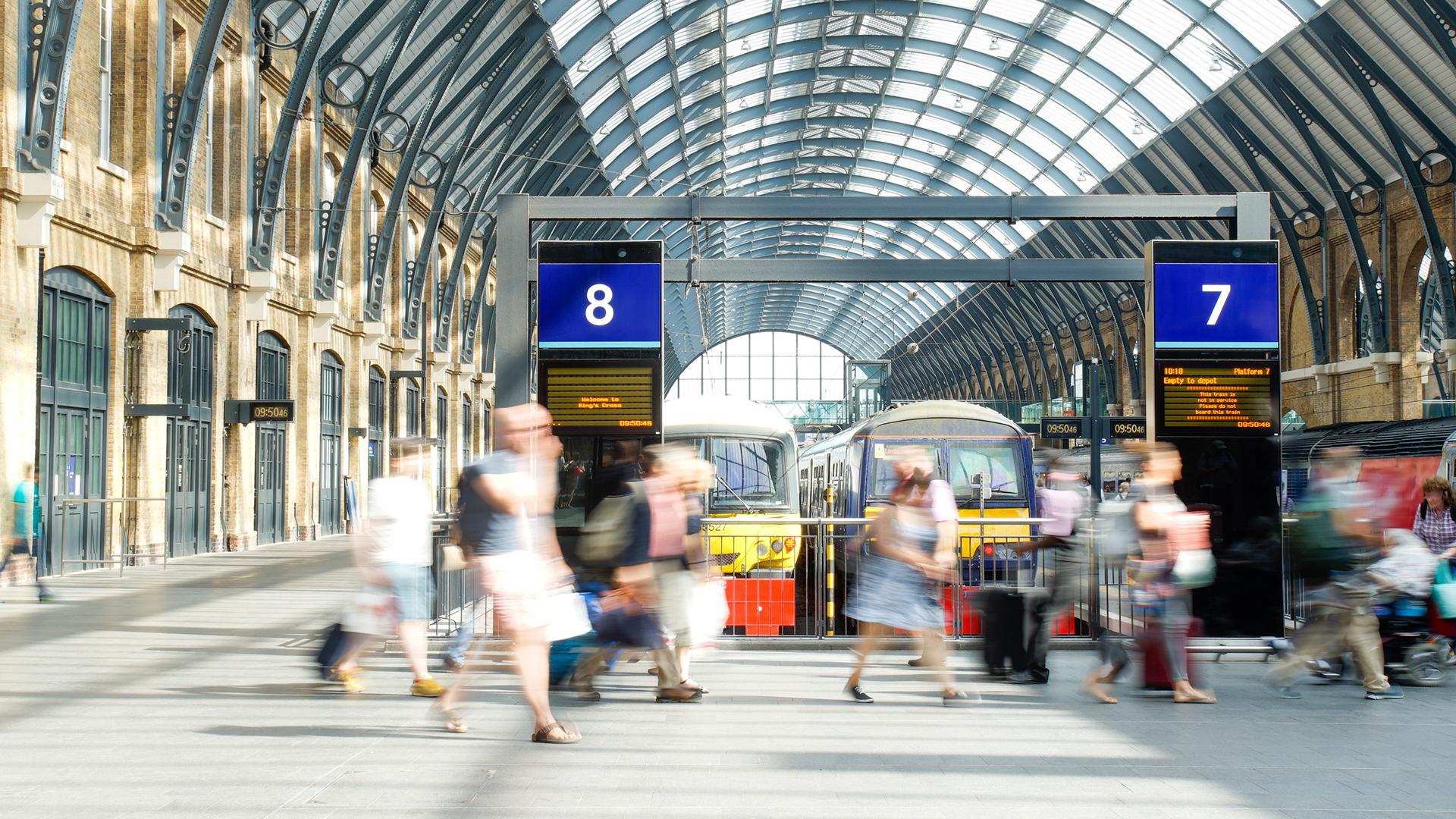 A busy train station with blurred motion of people walking, flanked by platforms 7 and 8, under a large glass arched roof.