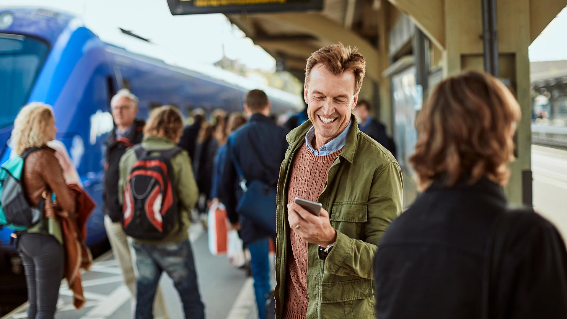 Man smiling at his phone on a busy train platform, surrounded by people and a blue train in the background.