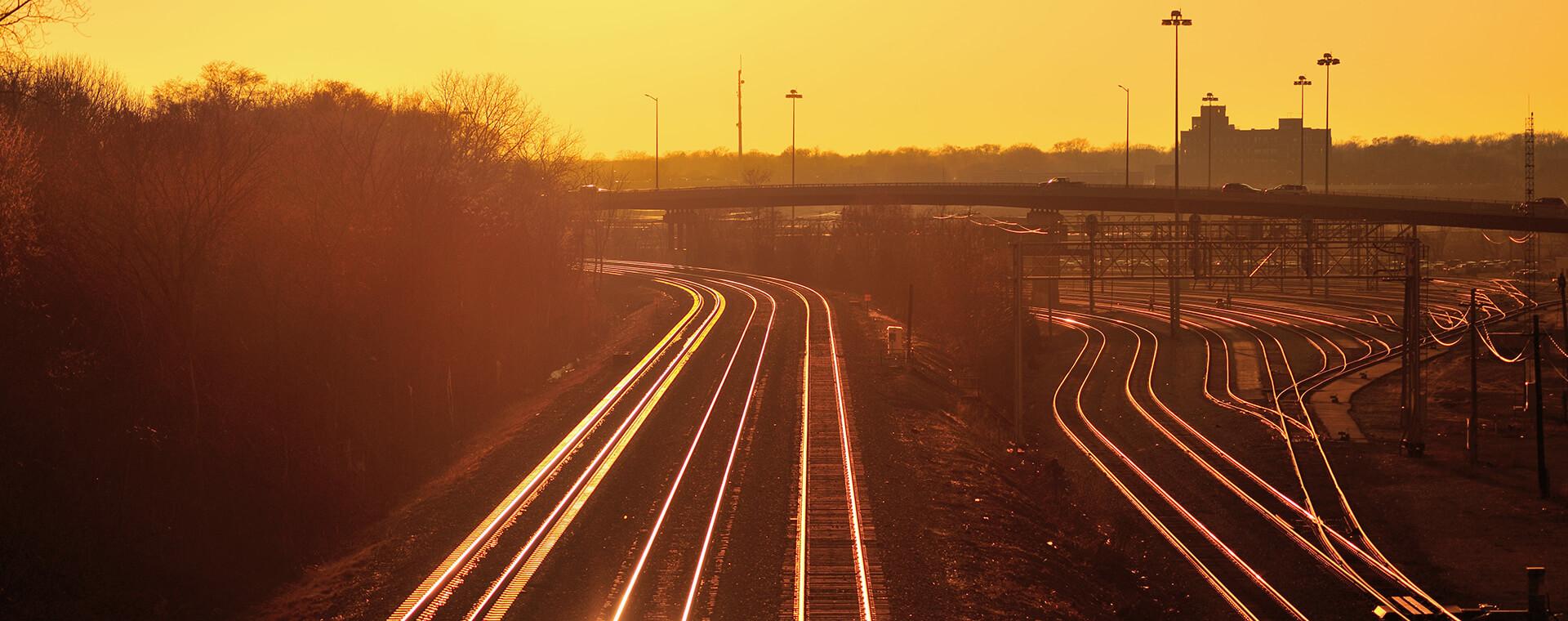 Railway tracks curve under a bridge during a golden sunset, with silhouettes of trees and distant buildings in the background.