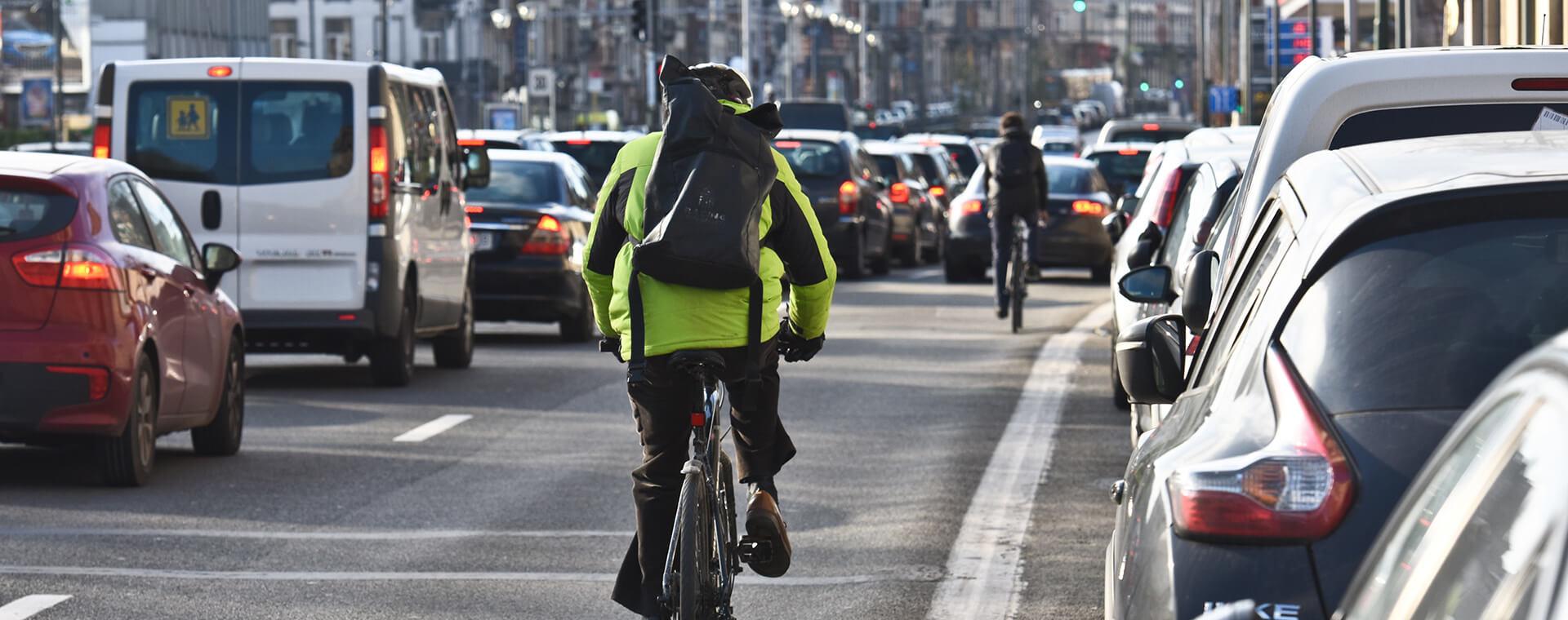 Cyclists ride between lanes of heavy traffic on a city street, with one wearing a bright green jacket and backpack.