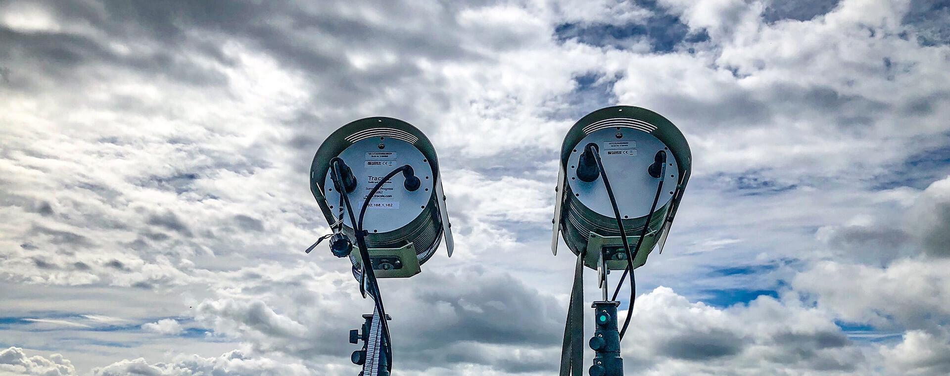 Two large outdoor spotlights facing upward against a cloudy sky, mounted on tall poles with visible cables and connectors.