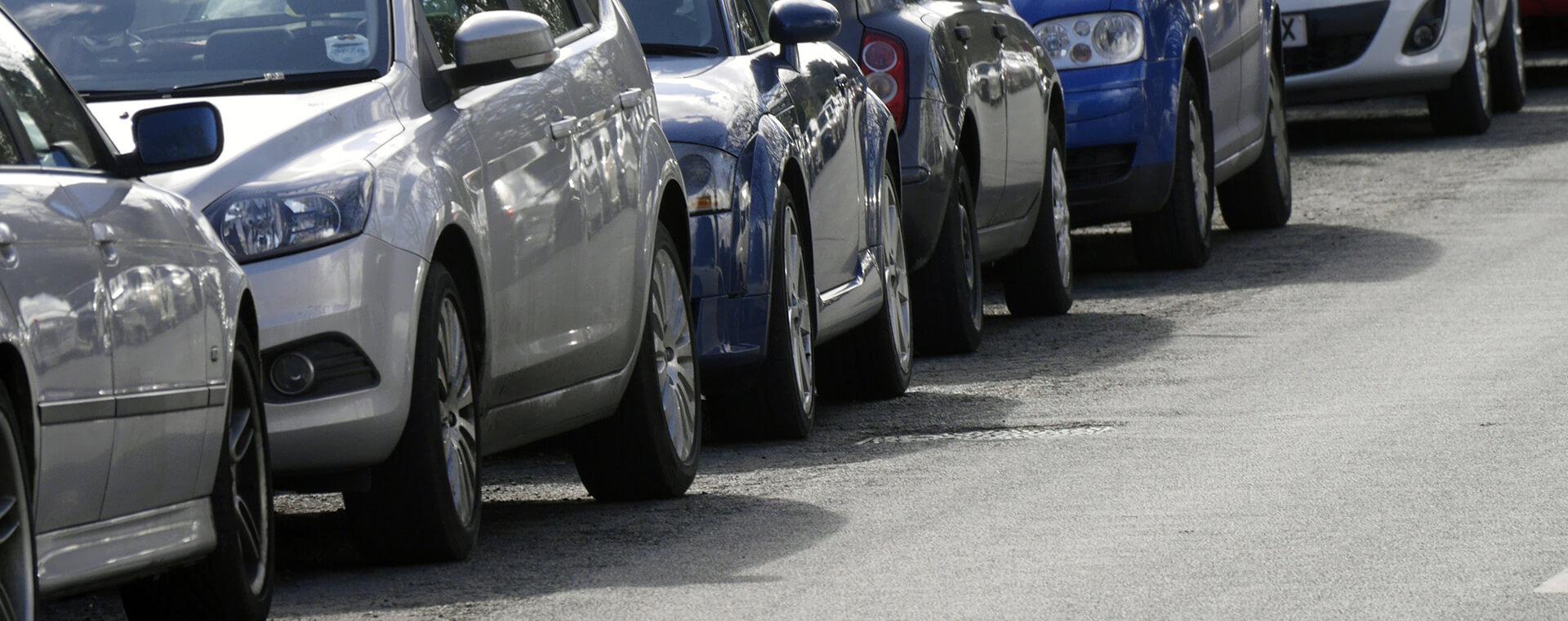 A row of parked cars in various colors lines a street, with the focus on the front ends and headlights.