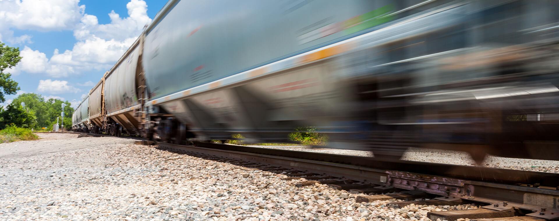 A fast-moving freight train blurs past on a sunny day, with a gravel track and green foliage in the background.