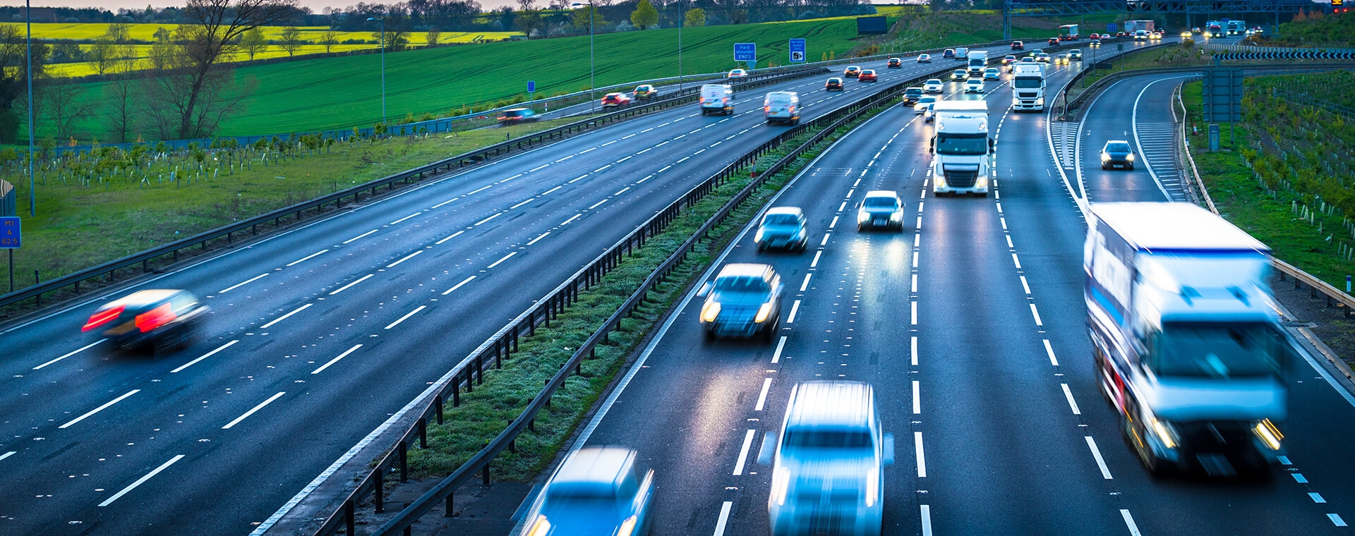 Busy highway with multiple lanes of fast-moving cars and trucks, bordered by green fields under a clear sky.