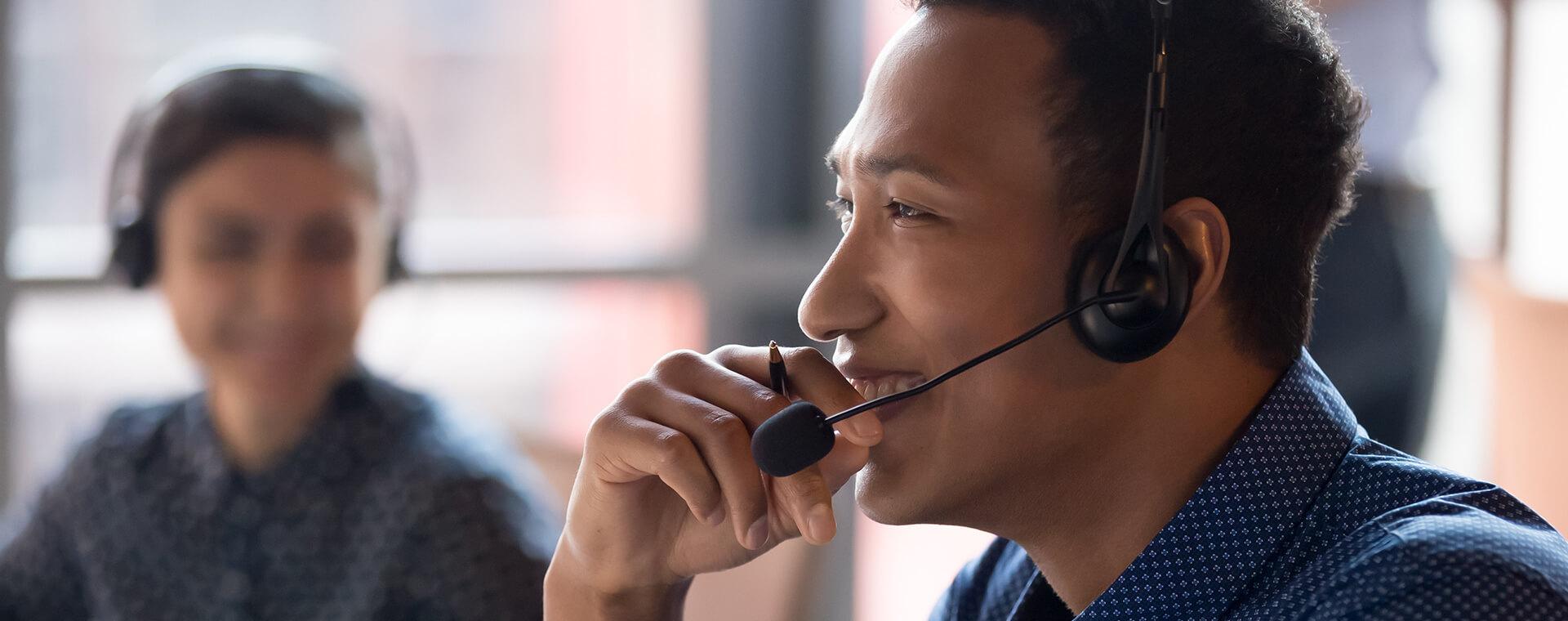 Man wearing a headset, smiling and talking, with a blurred colleague in the background, both in a bright office setting.