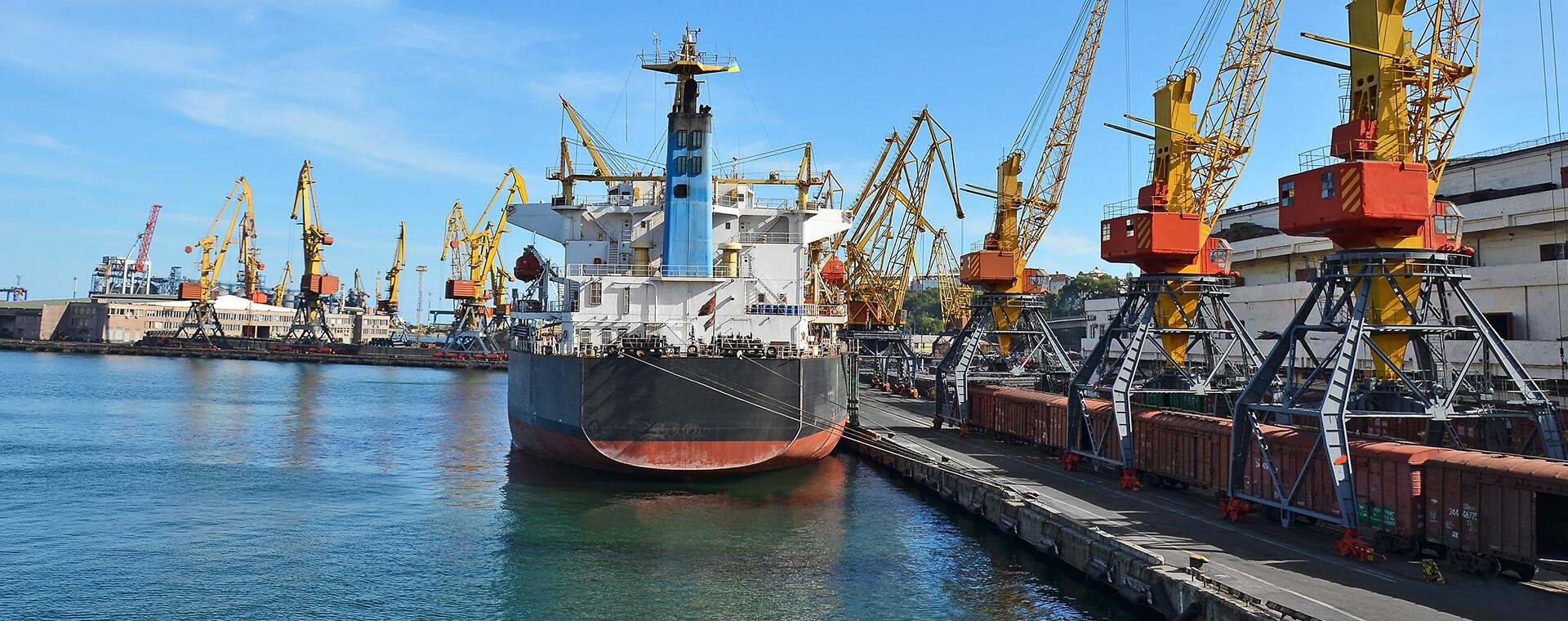 A large cargo ship docked at an industrial port, surrounded by numerous yellow cranes and shipping containers under a clear blue sky.