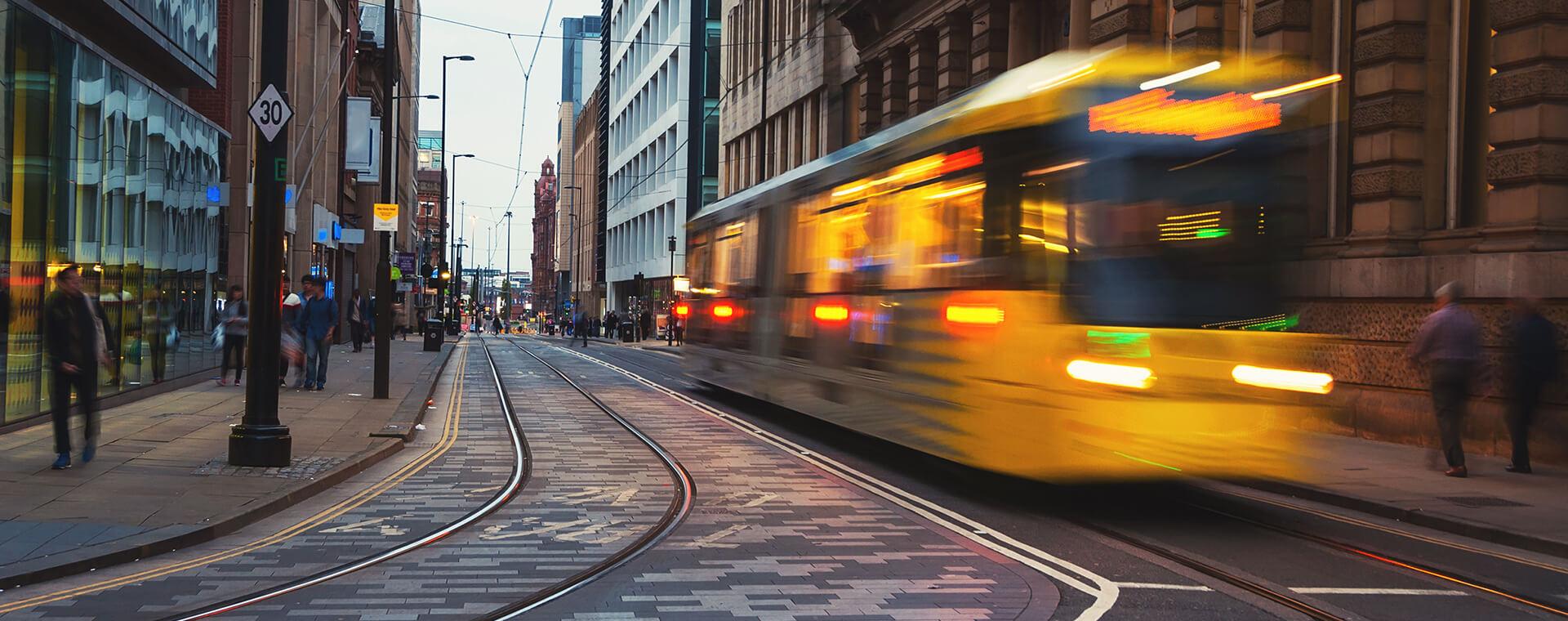 A yellow tram speeds through a city street, surrounded by modern and historic buildings, with pedestrians on the sidewalk.