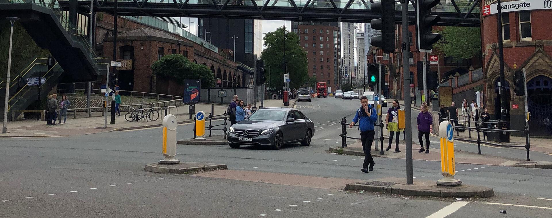 A city street intersection with pedestrians crossing, a black car turning, and buildings in the background. Traffic lights are visible.