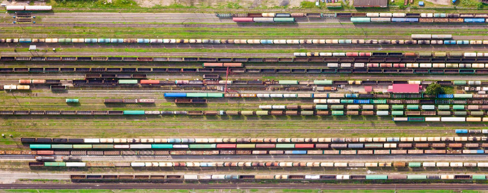 Aerial view of a railway yard with numerous colorful freight train cars on multiple parallel tracks, surrounded by greenery.