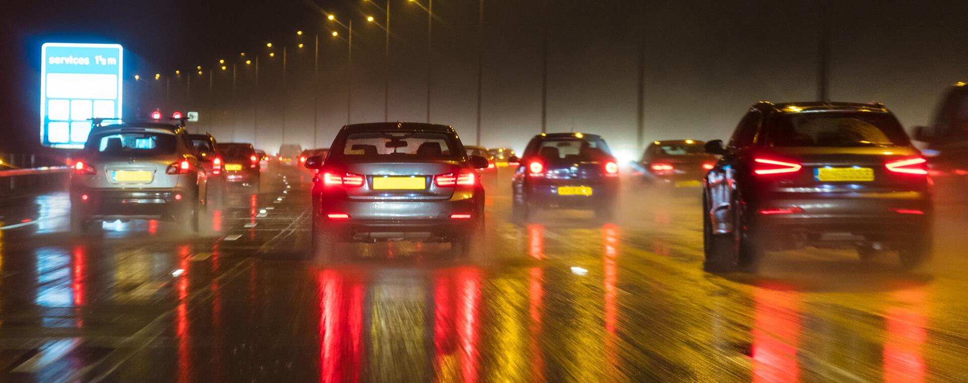 Cars driving on a wet highway at night, with bright headlights and reflections on the road, under streetlights.