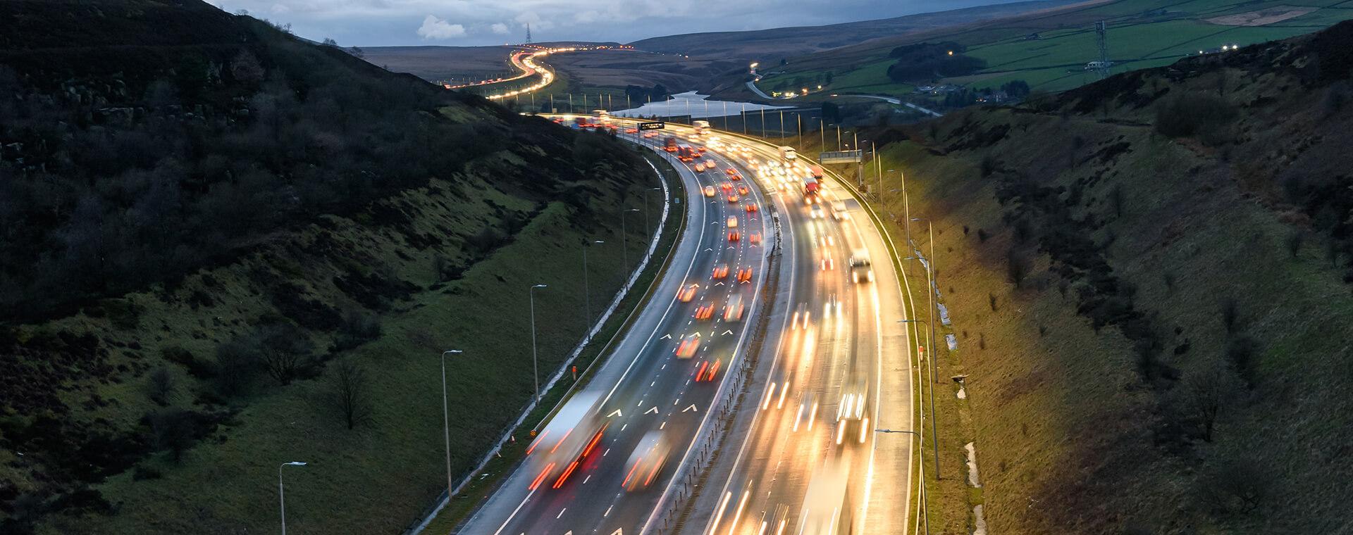 Aerial view of a busy highway at dusk with blurred car lights, surrounded by hills and fields under a cloudy sky.