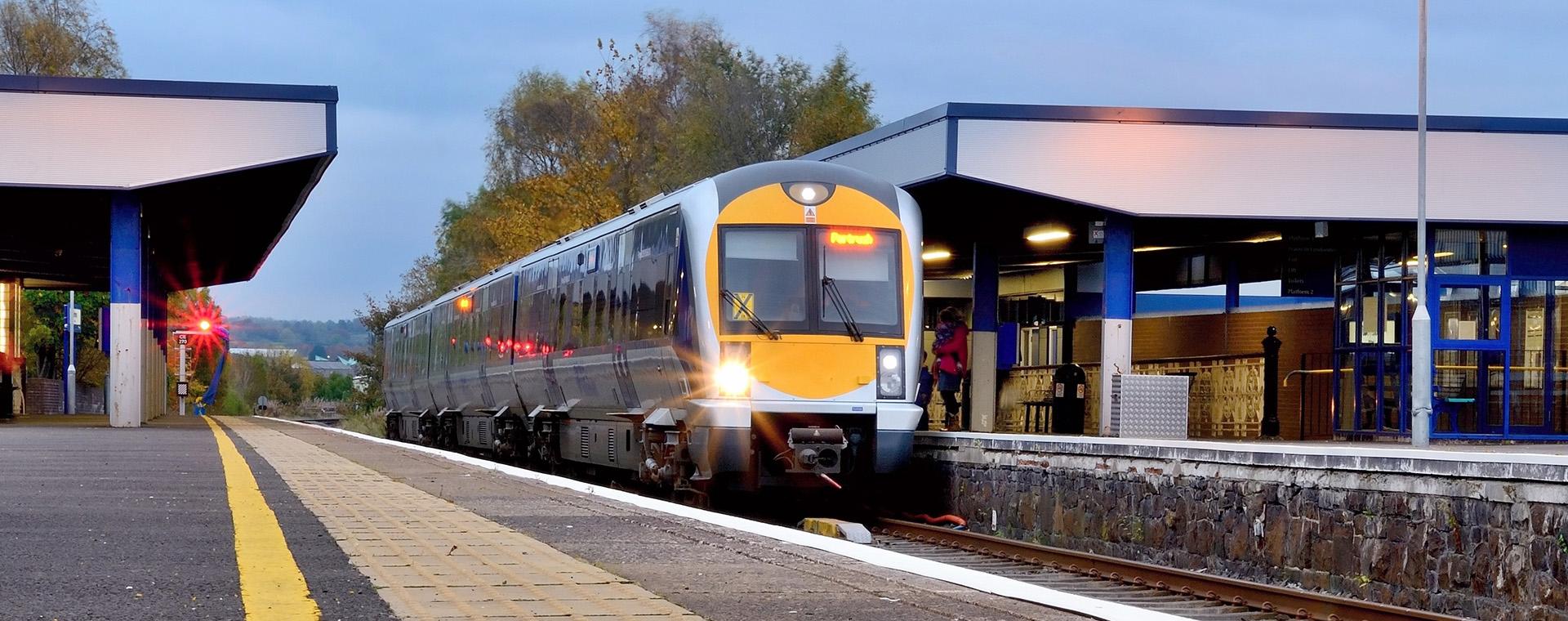A modern train arrives at a quiet station platform under a cloudy sky, with trees in the background and platform lights glowing.