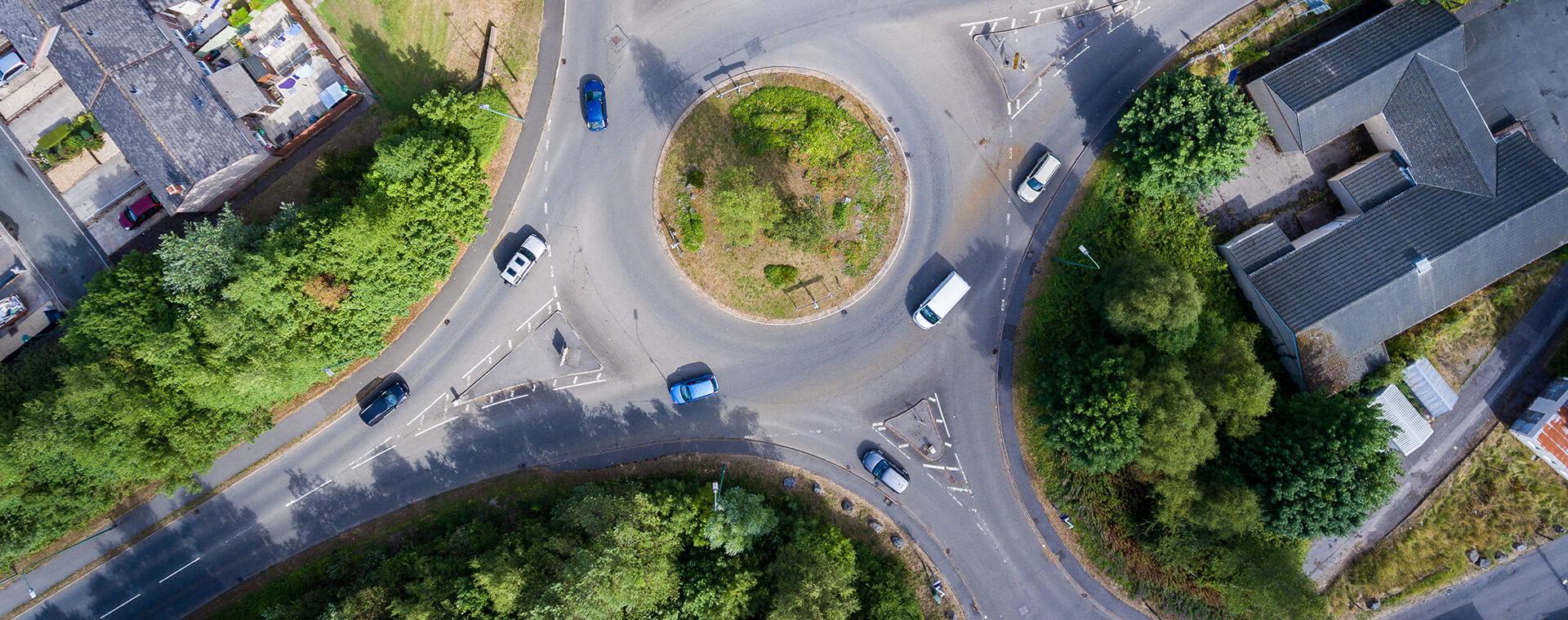 Aerial view of a roundabout with several cars, surrounded by greenery and buildings.