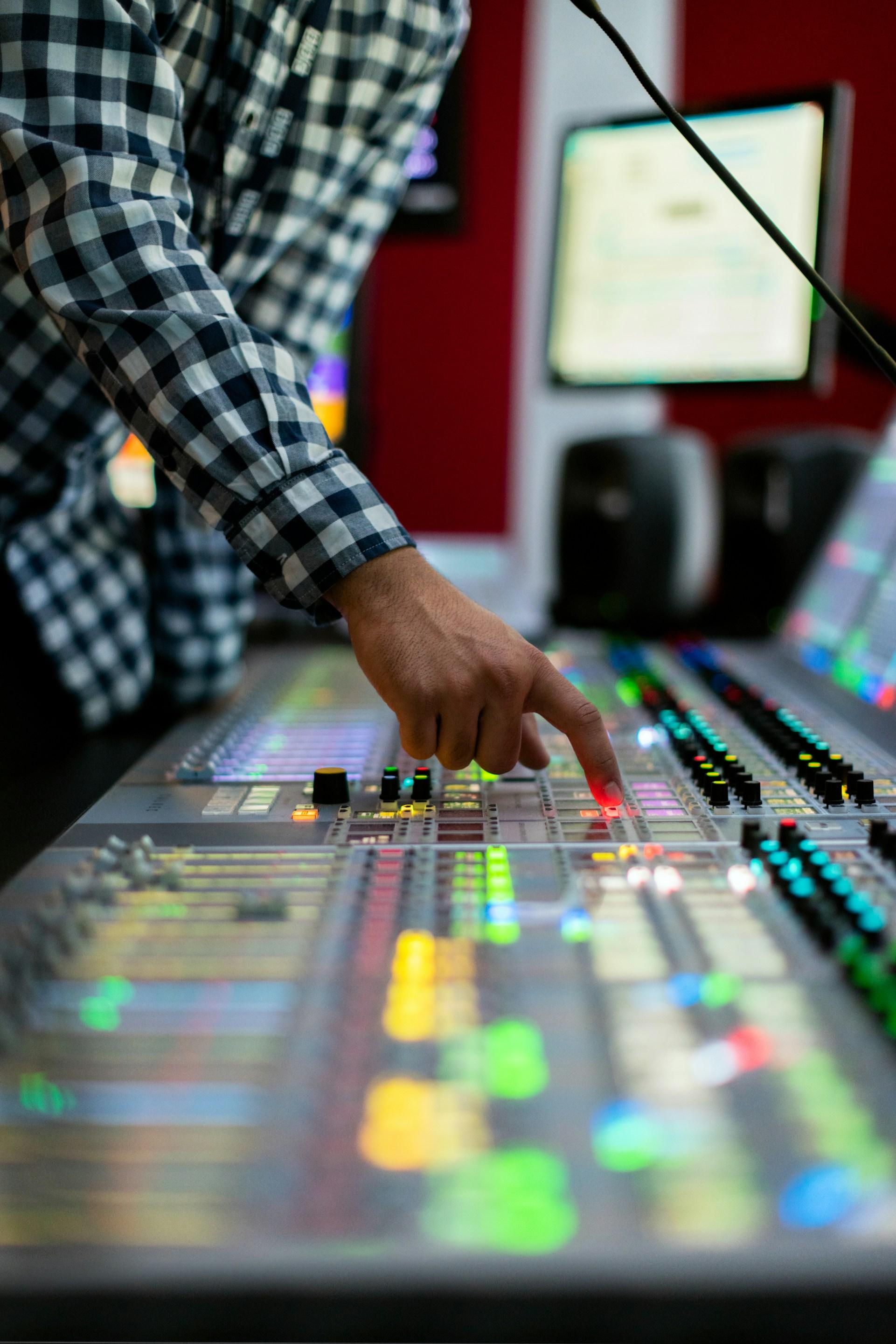Person adjusting controls on a brightly lit audio mixing console in a studio, wearing a checkered shirt.