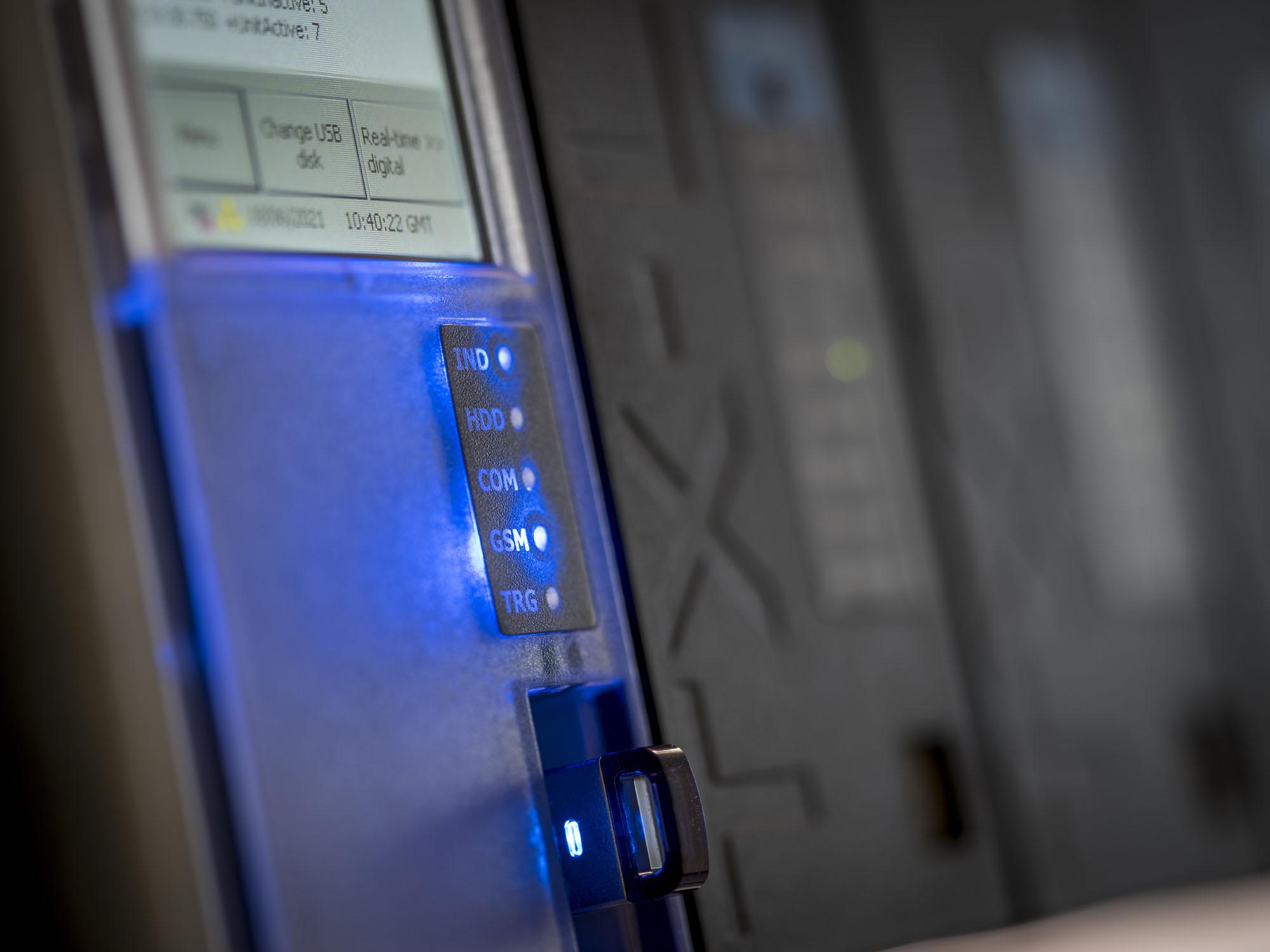 Close-up of a digital control panel with illuminated blue indicators labeled IND, HDD, COM, GSM, and TRG, with a USB device inserted.
