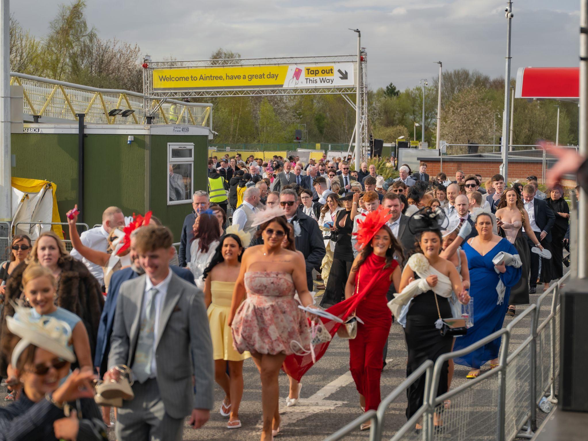 A large crowd of people dressed in formal attire walk under a "Welcome to Aintree" sign, attending an outdoor event.