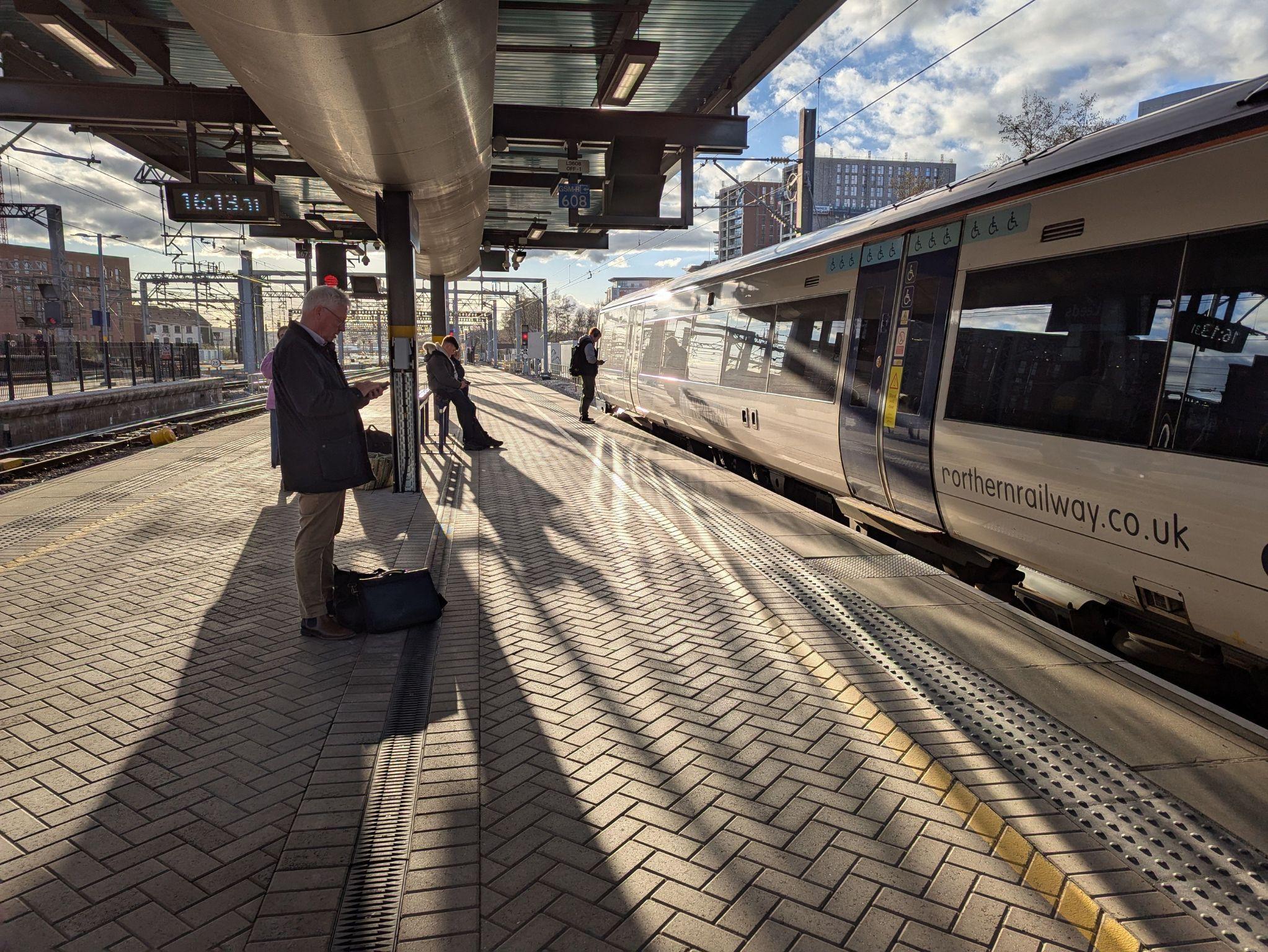 Commuters standing on Leeds railway platform with sunlit train and city skyline in background.