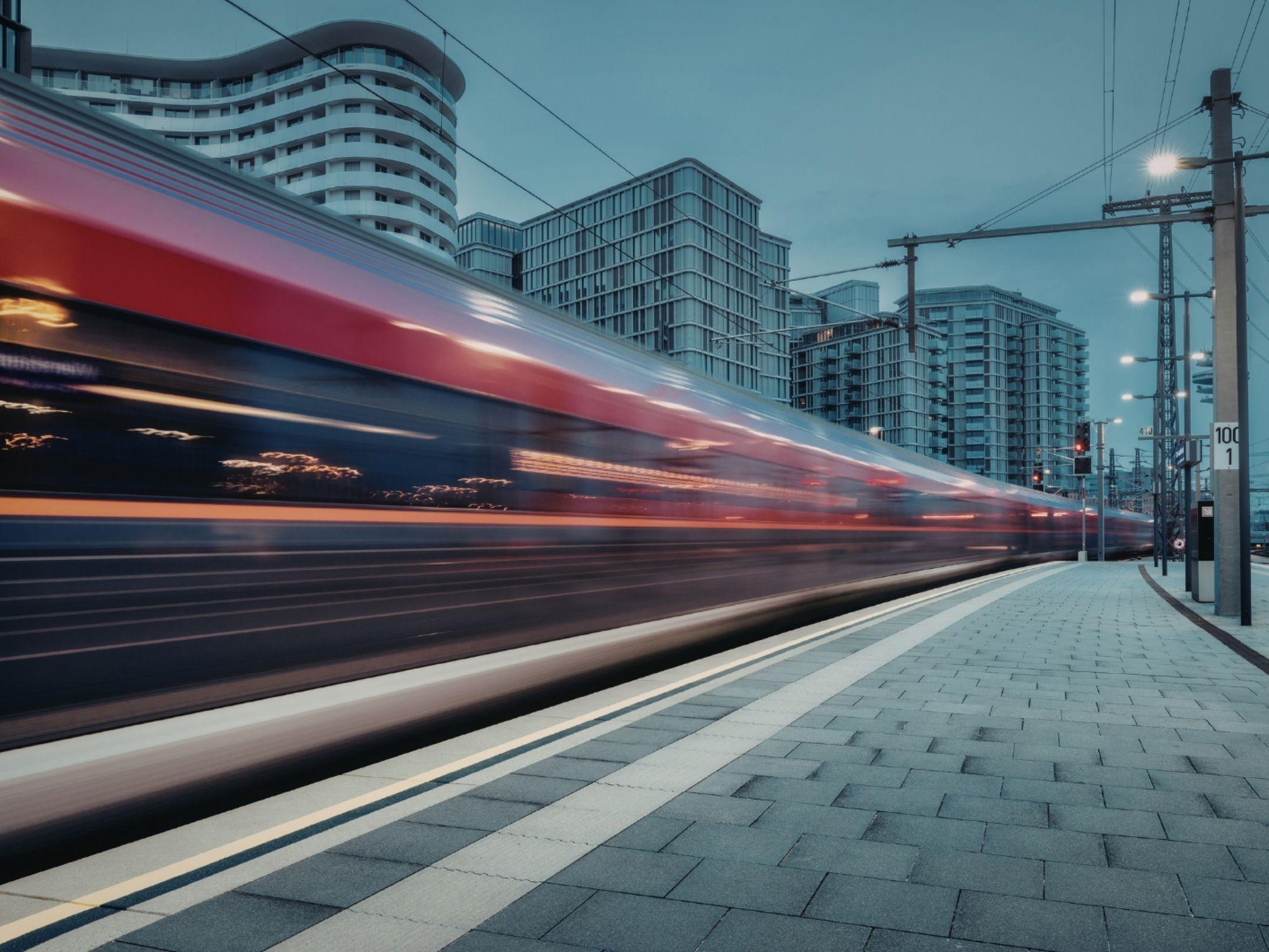 A red train blurs by at a modern urban station, surrounded by sleek high-rise buildings under a cloudy sky.