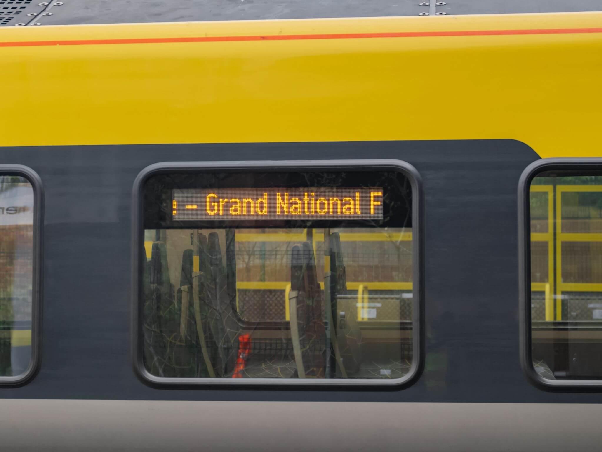 Yellow train with a digital display in the window reading "Grand National F," viewed from the side.