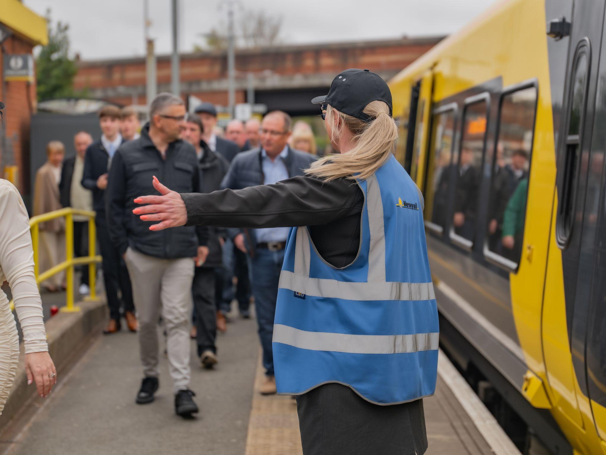 Rail worker in a blue vest directs passengers at a train station. A yellow train is alongside the platform, and people are walking by.